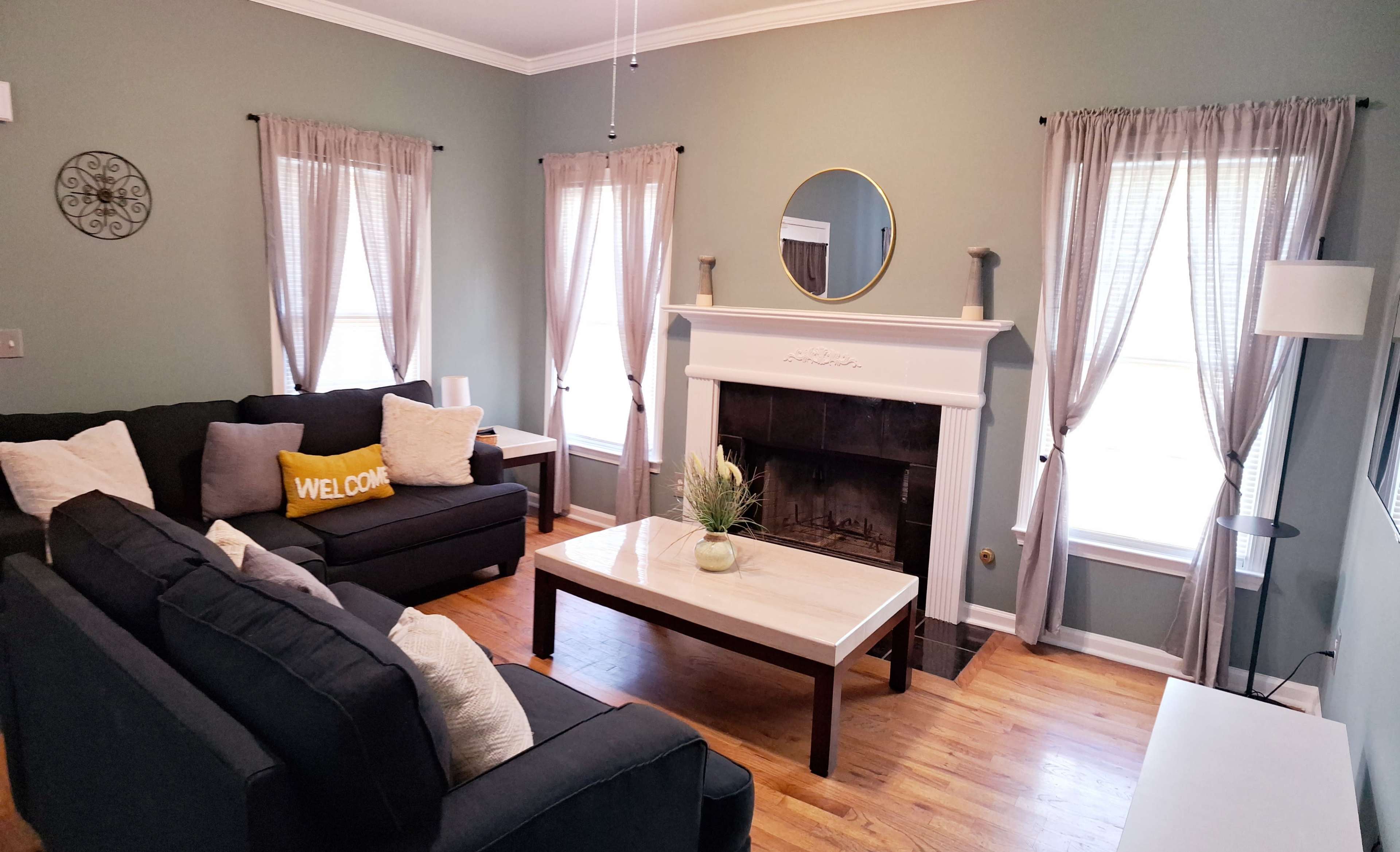 The living room features a dark sofa set, a white marble coffee table, and a fireplace with a round mirror above it, surrounded by light curtains and wooden flooring.