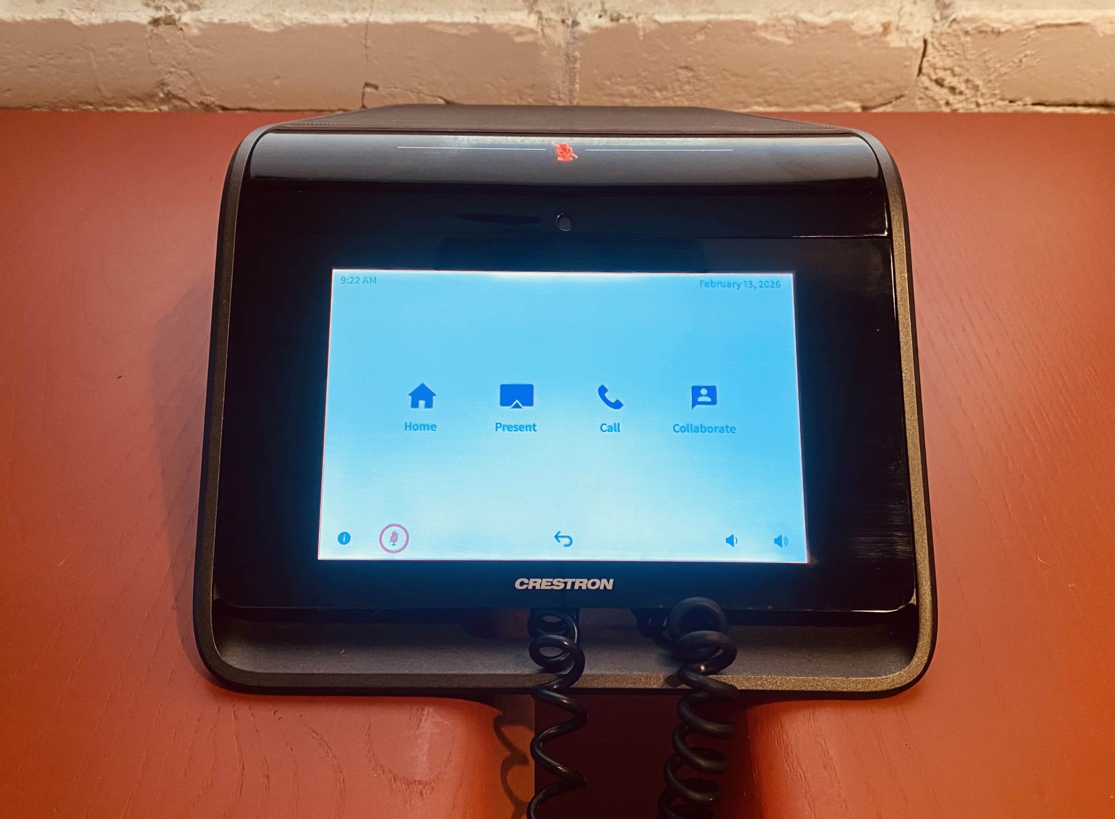 The image shows a Crestron touchscreen interface mounted on a wall, displaying options for home, present, call, and collaborate, with a coiled phone handset connected.