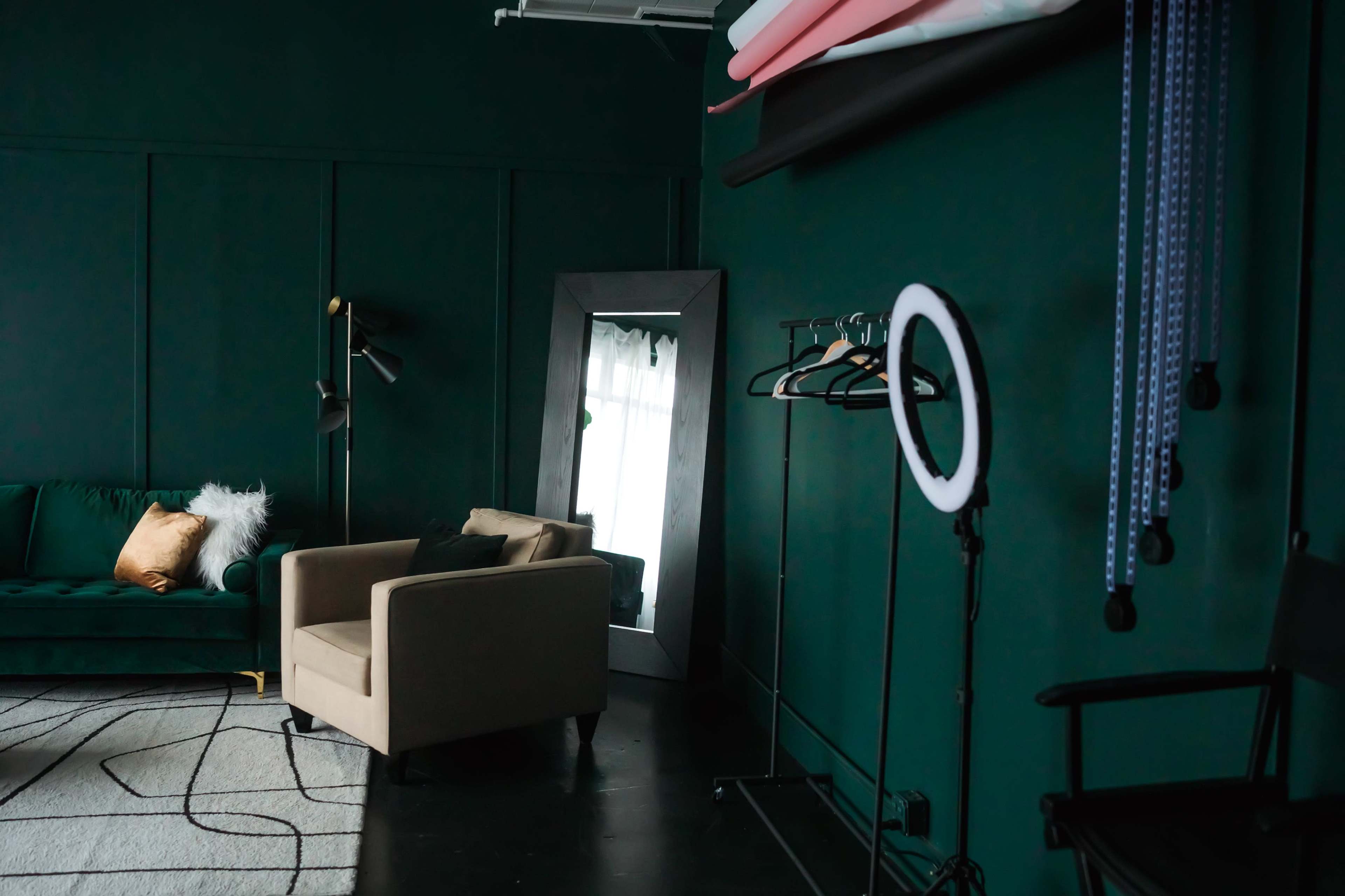 The image depicts a stylish interior with a dark green wall, a green sofa, a large mirror, a clothing rack, and a ring light.