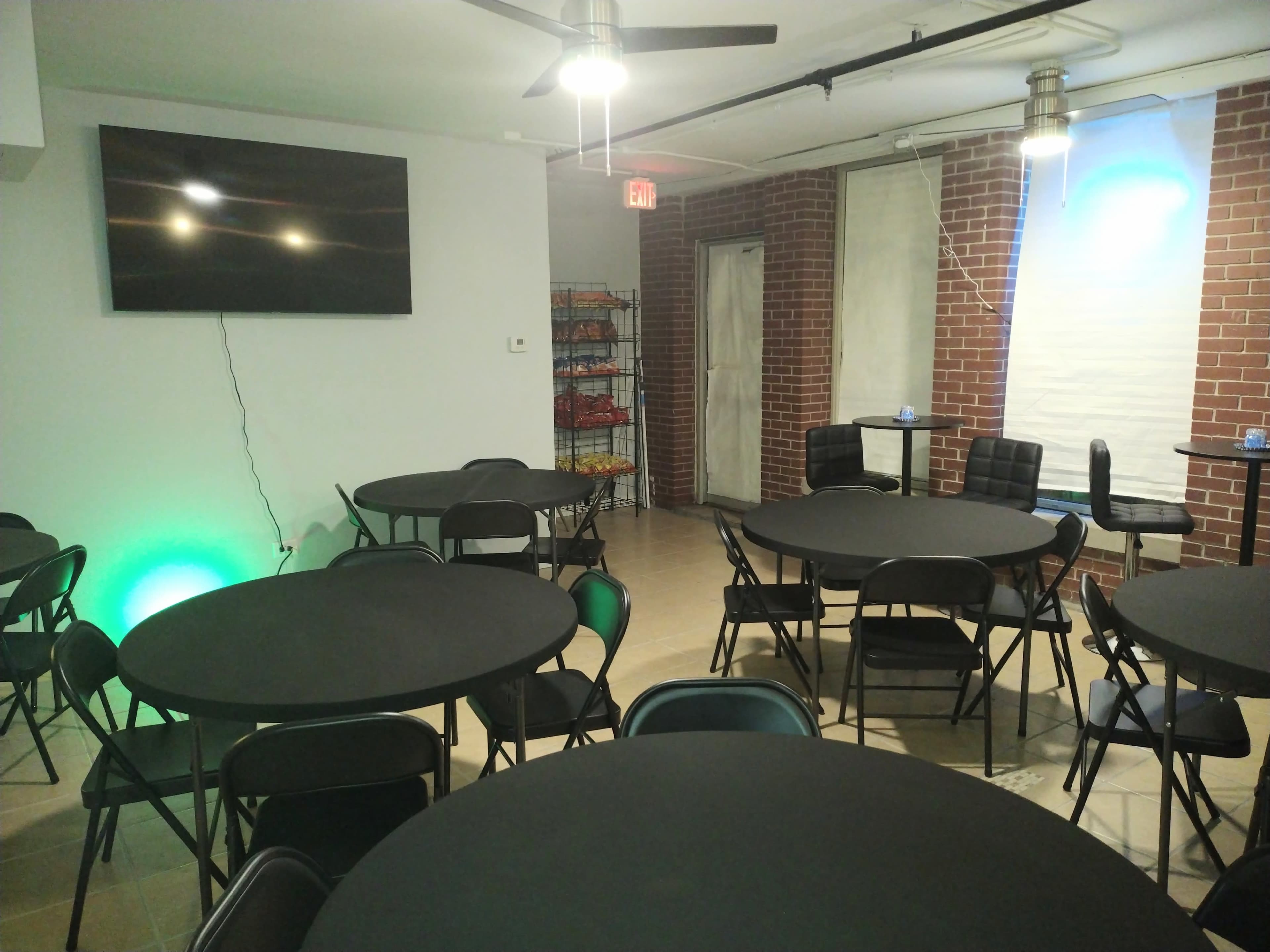 A meeting room with several round black tables and chairs arranged neatly, a television mounted on the wall, and a brick backdrop.