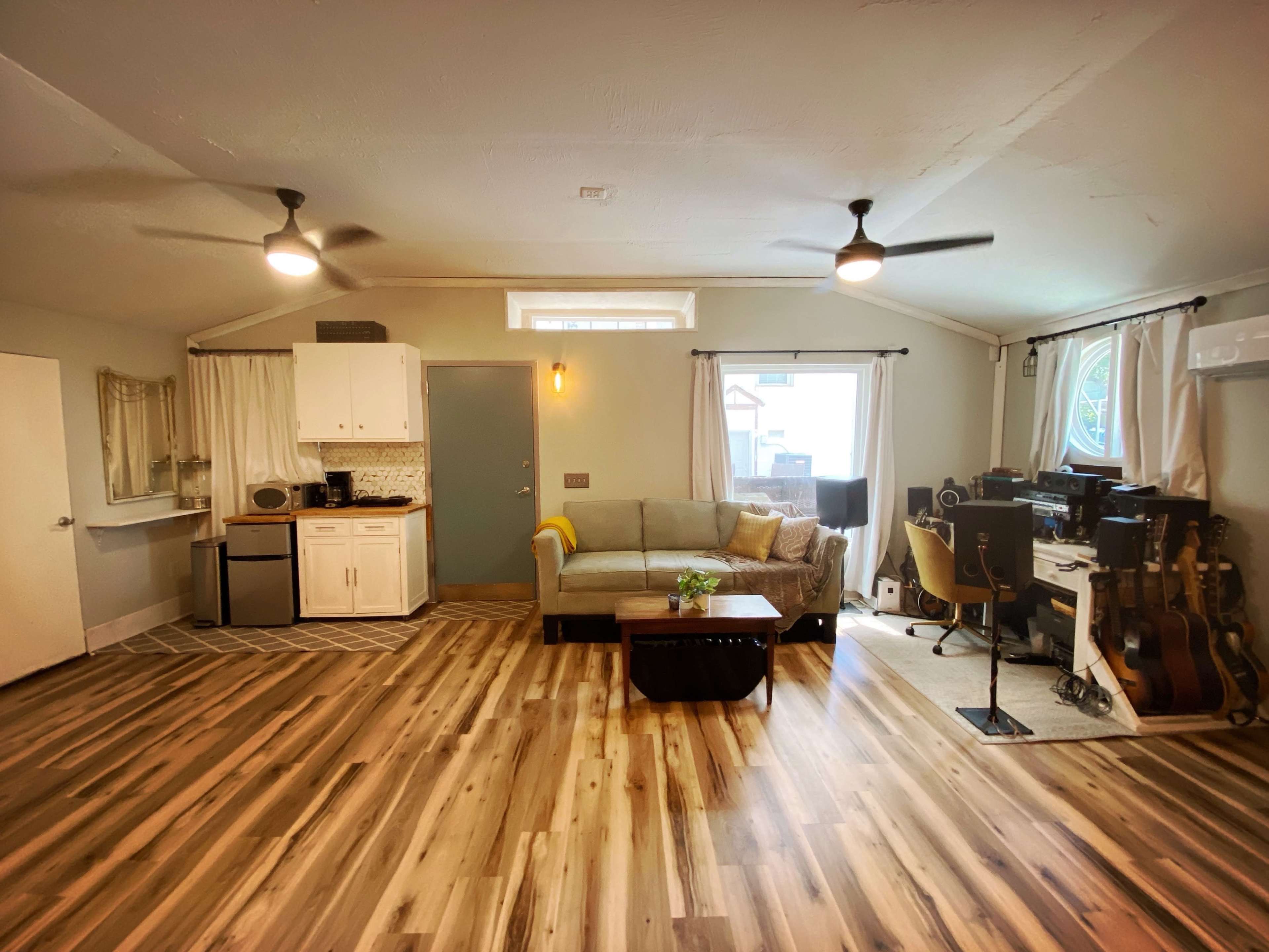 The image shows an open-concept living area with a kitchen, a sofa, and a workspace featuring musical instruments.