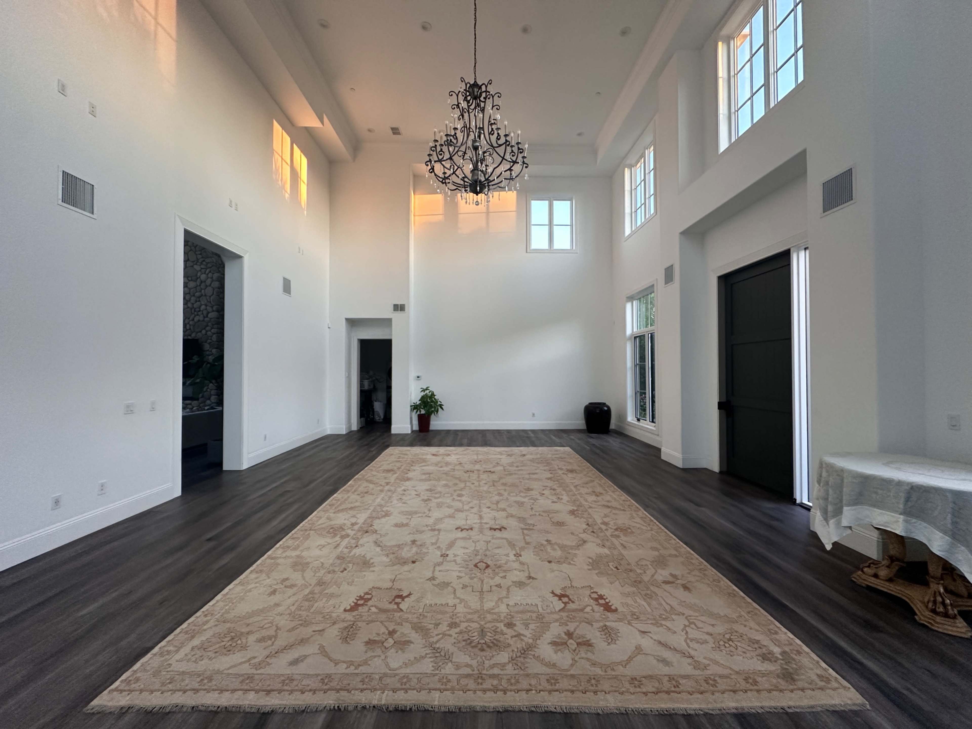 The image shows a spacious, well-lit room with high ceilings, featuring a chandelier, a large area rug, and windows that allow natural light to enter.