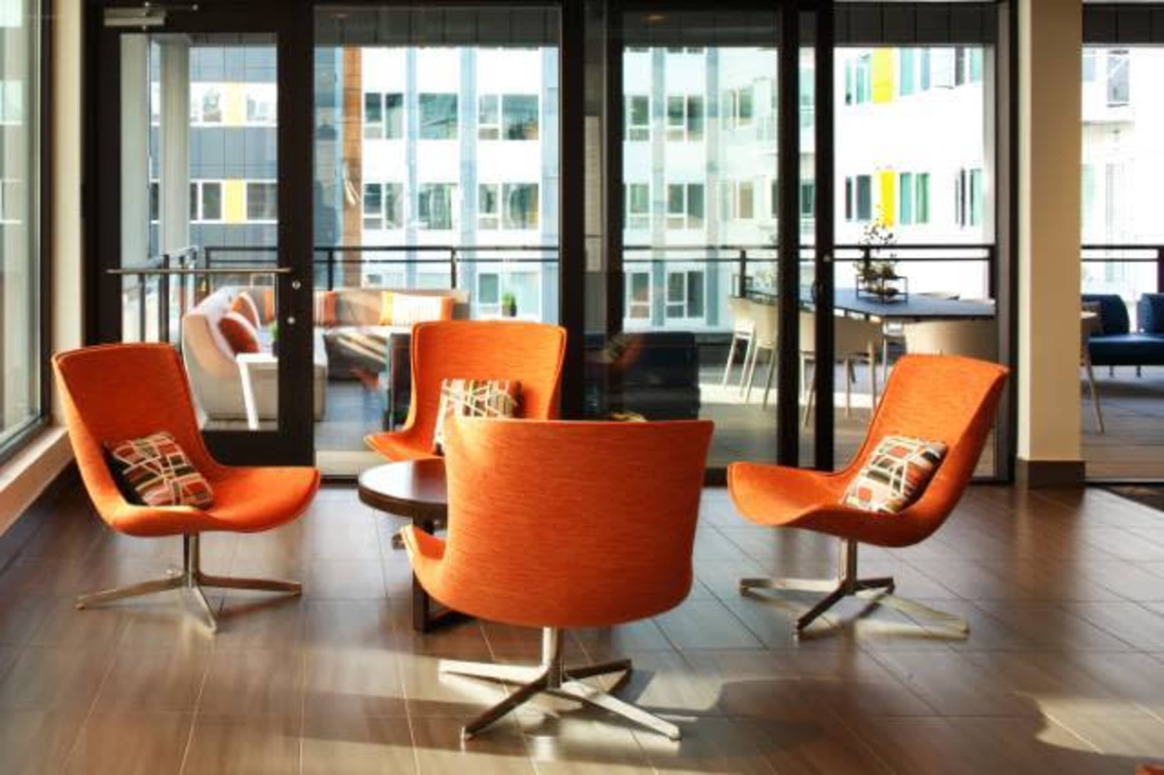 A modern lounge area with four orange chairs arranged around a circular table, surrounded by large glass windows.