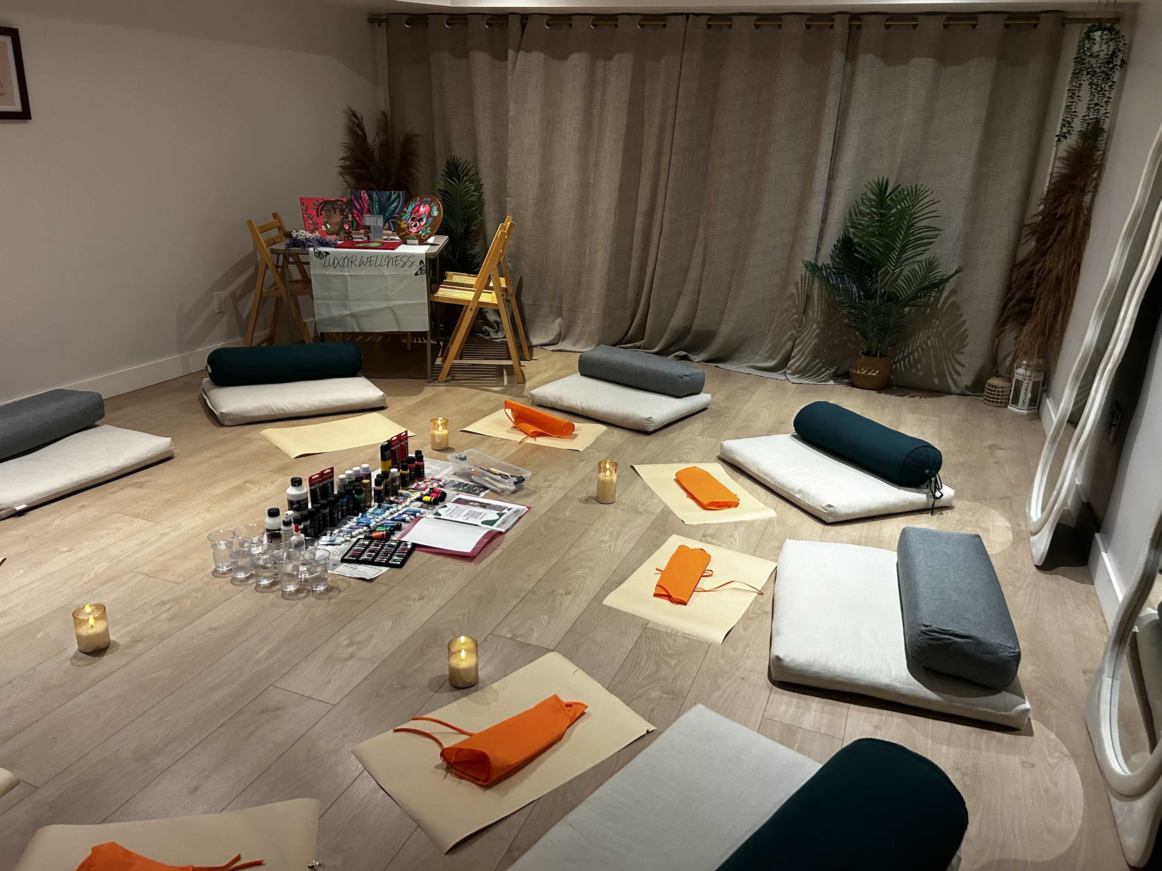 The image shows a neatly arranged meditation or workshop space with cushions on the floor, candles, craft supplies, and a table set in the background.
