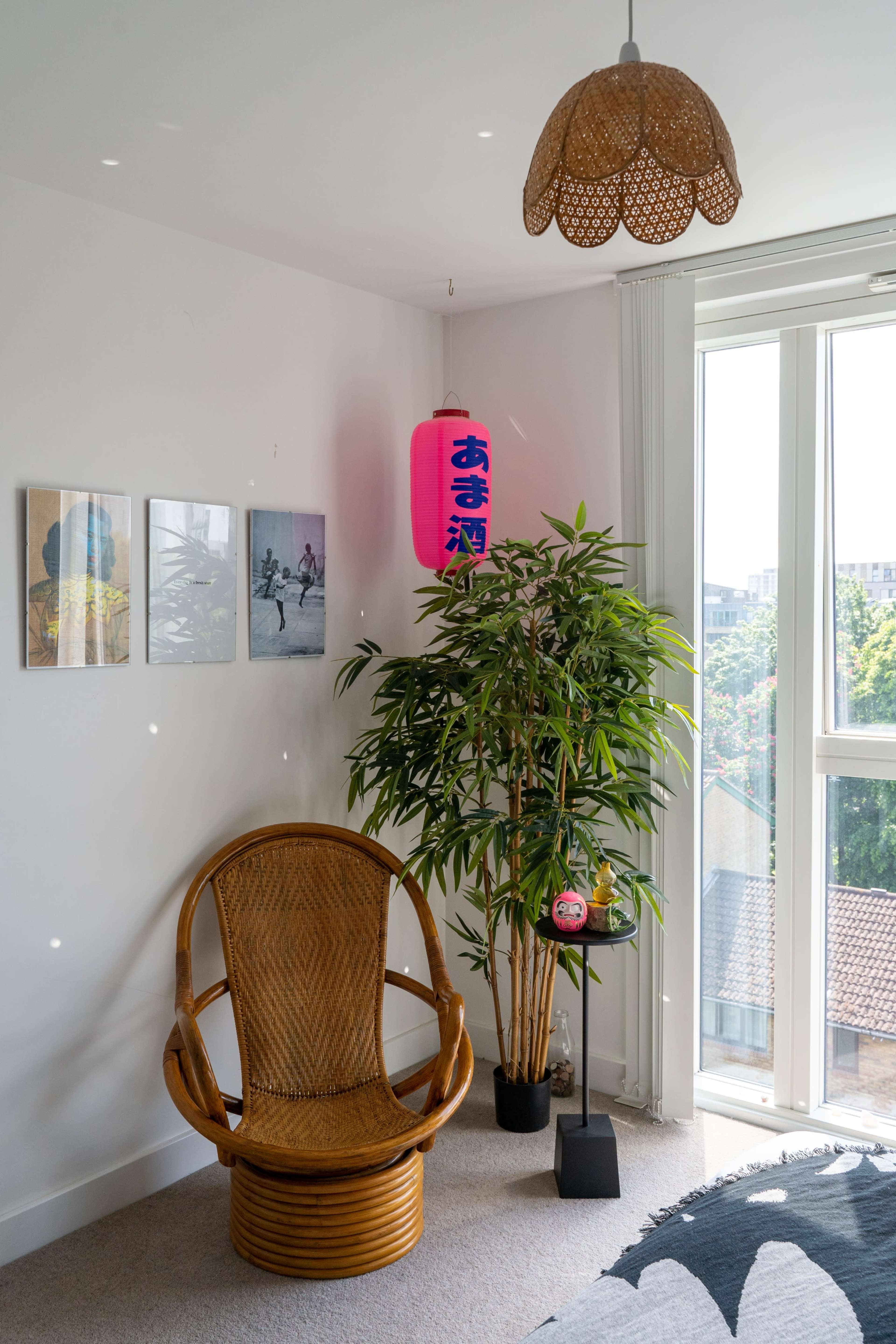 A corner of a room with a rattan chair, a potted bamboo plant, a colorful lantern hanging from the ceiling, and framed photos on the wall.