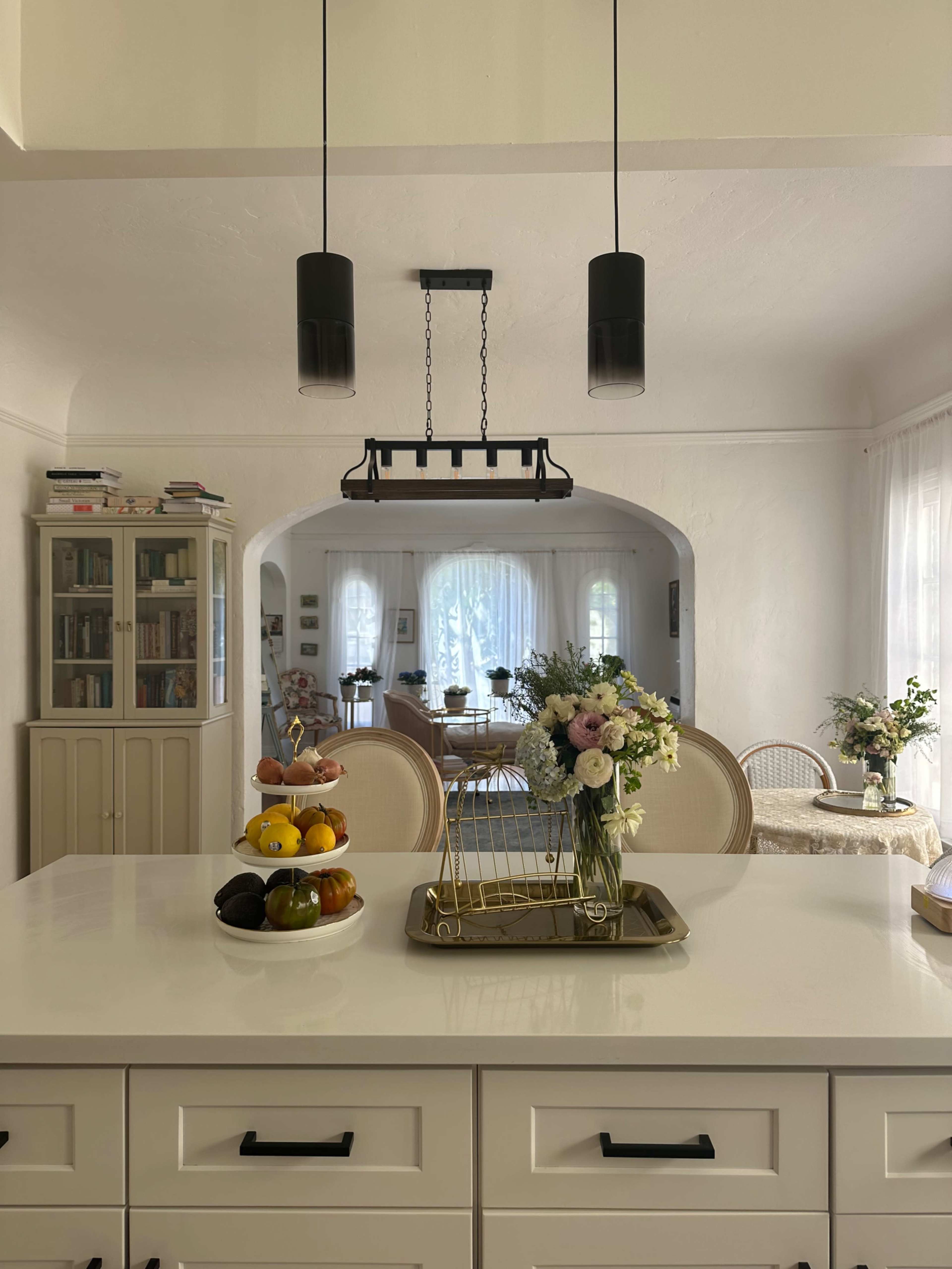 A modern kitchen features a central island with a fruit basket, a floral arrangement, and pendant lighting, overlooking a cozy dining area through an archway.