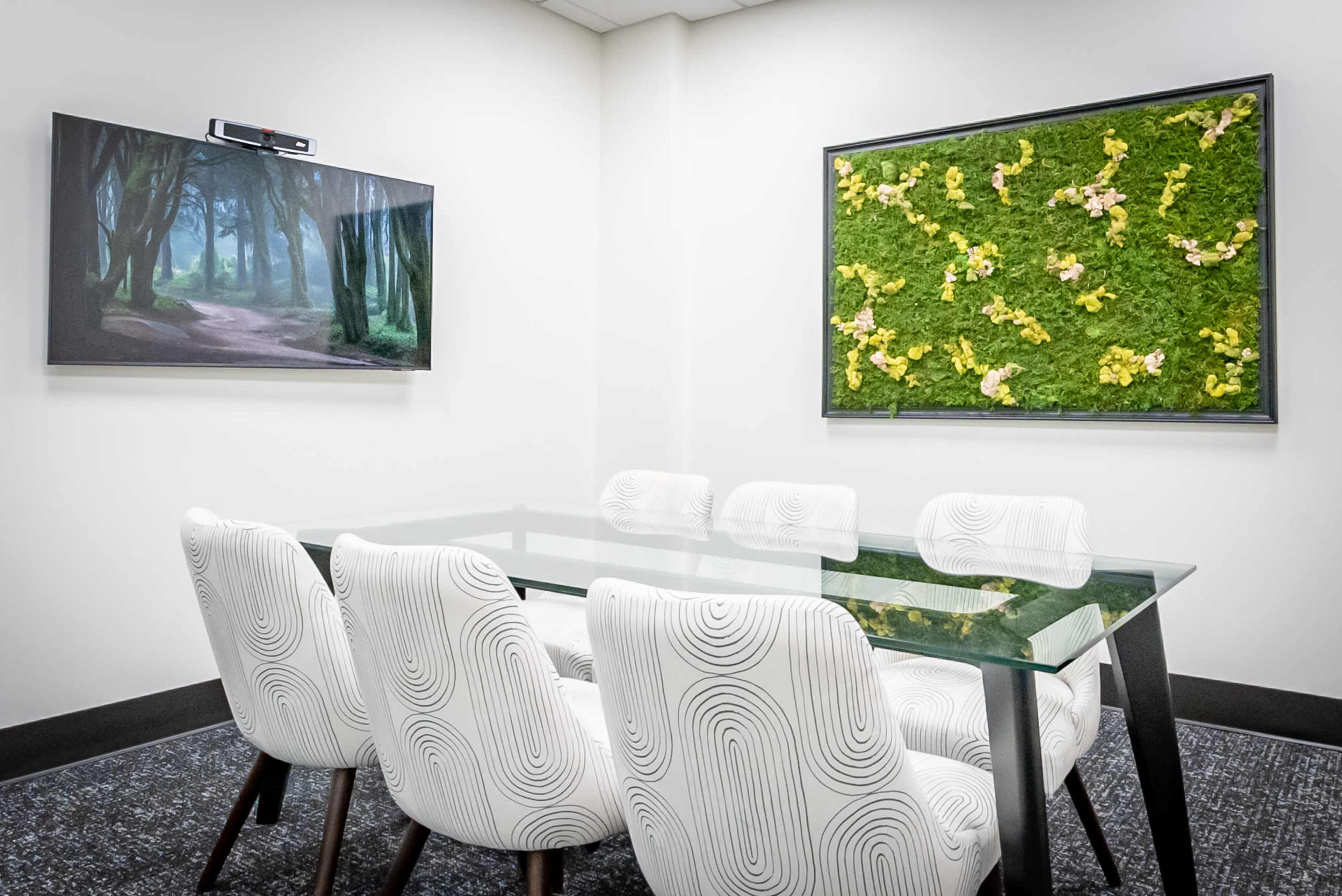 The image shows a modern conference room featuring a glass table surrounded by white chairs, with a nature-themed television and a green wall art piece.