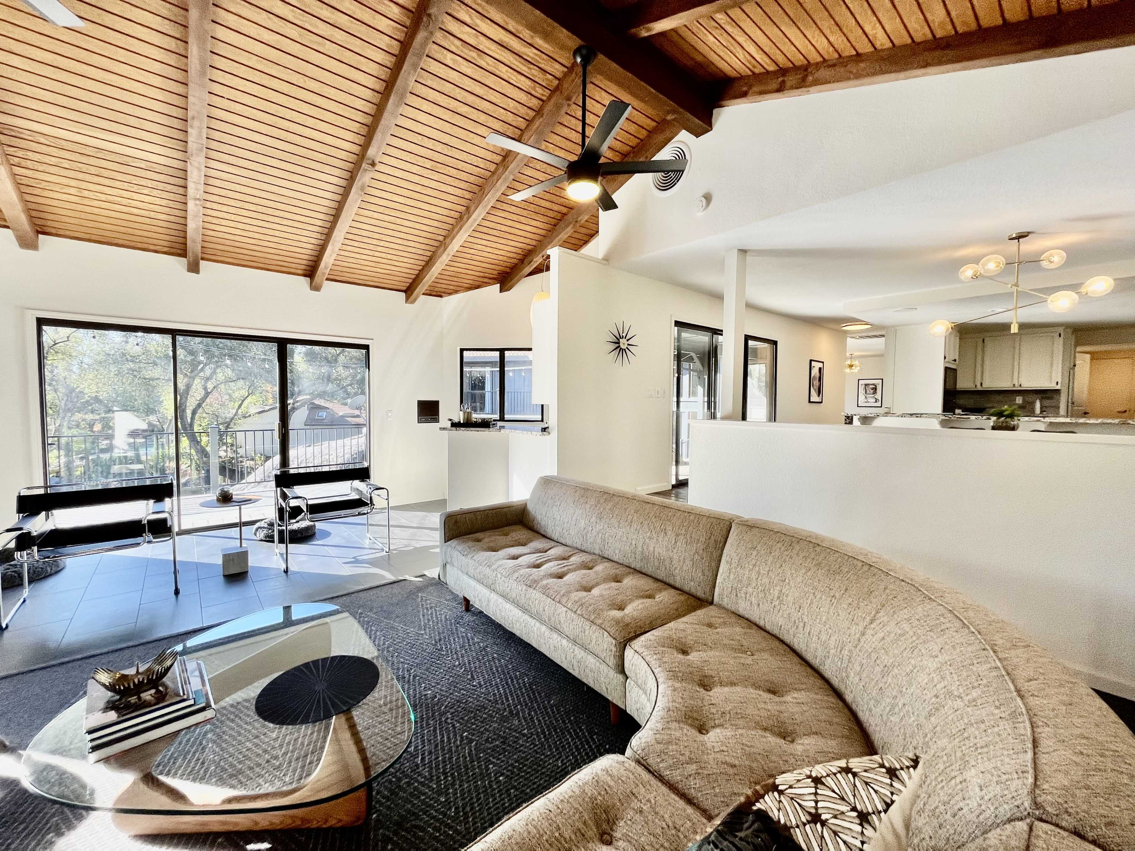 A modern living room features a curved sectional sofa, a glass coffee table, and large windows that provide natural light, all set under a vaulted wooden ceiling.