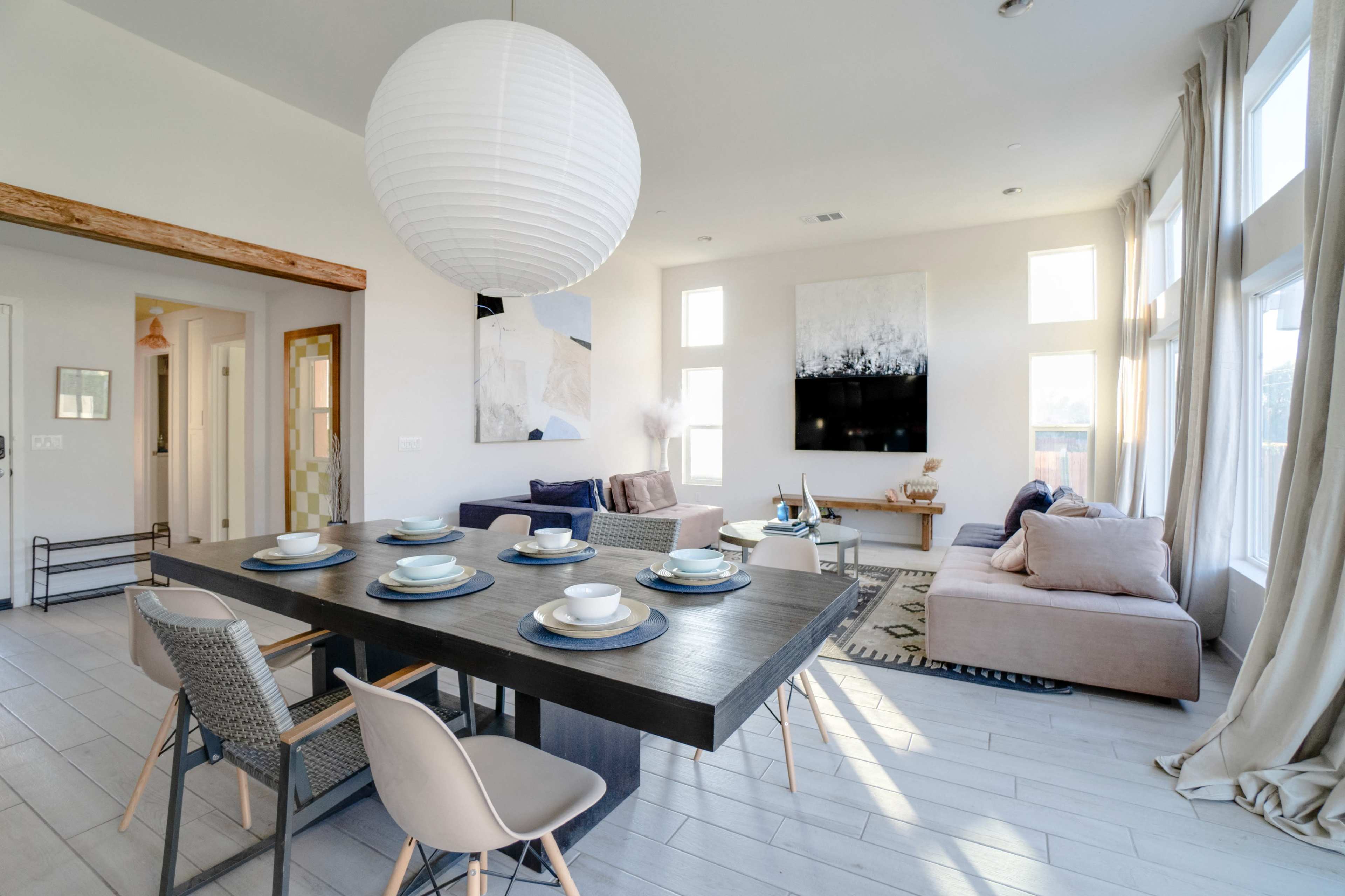 A modern living and dining area featuring a table set for six, a large wall-mounted television, and various seating arrangements with natural light flooding through large windows.