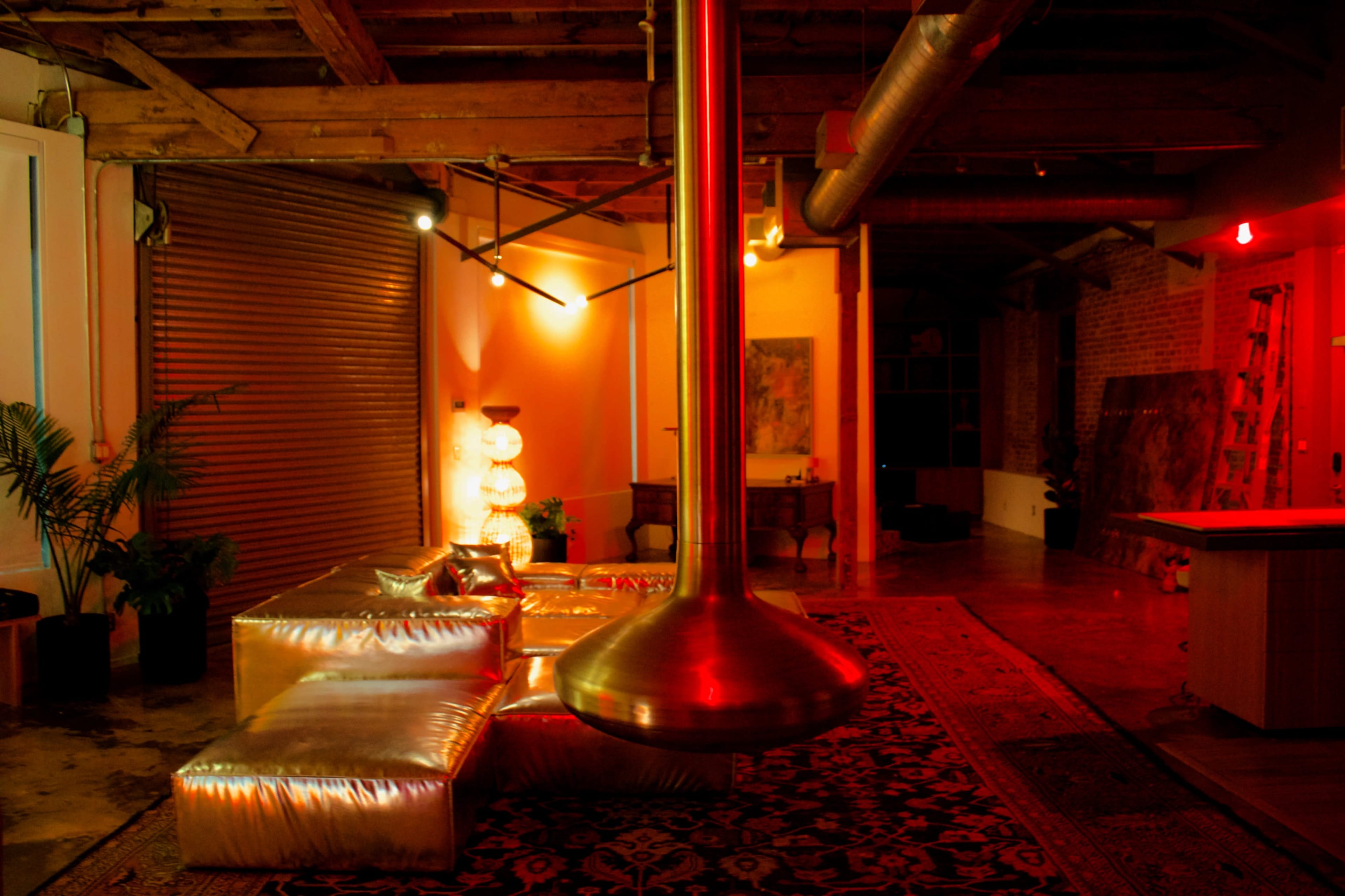 The image shows an industrial-style interior with metallic furniture, a prominent hanging light fixture, and a rug under soft, colorful lighting.