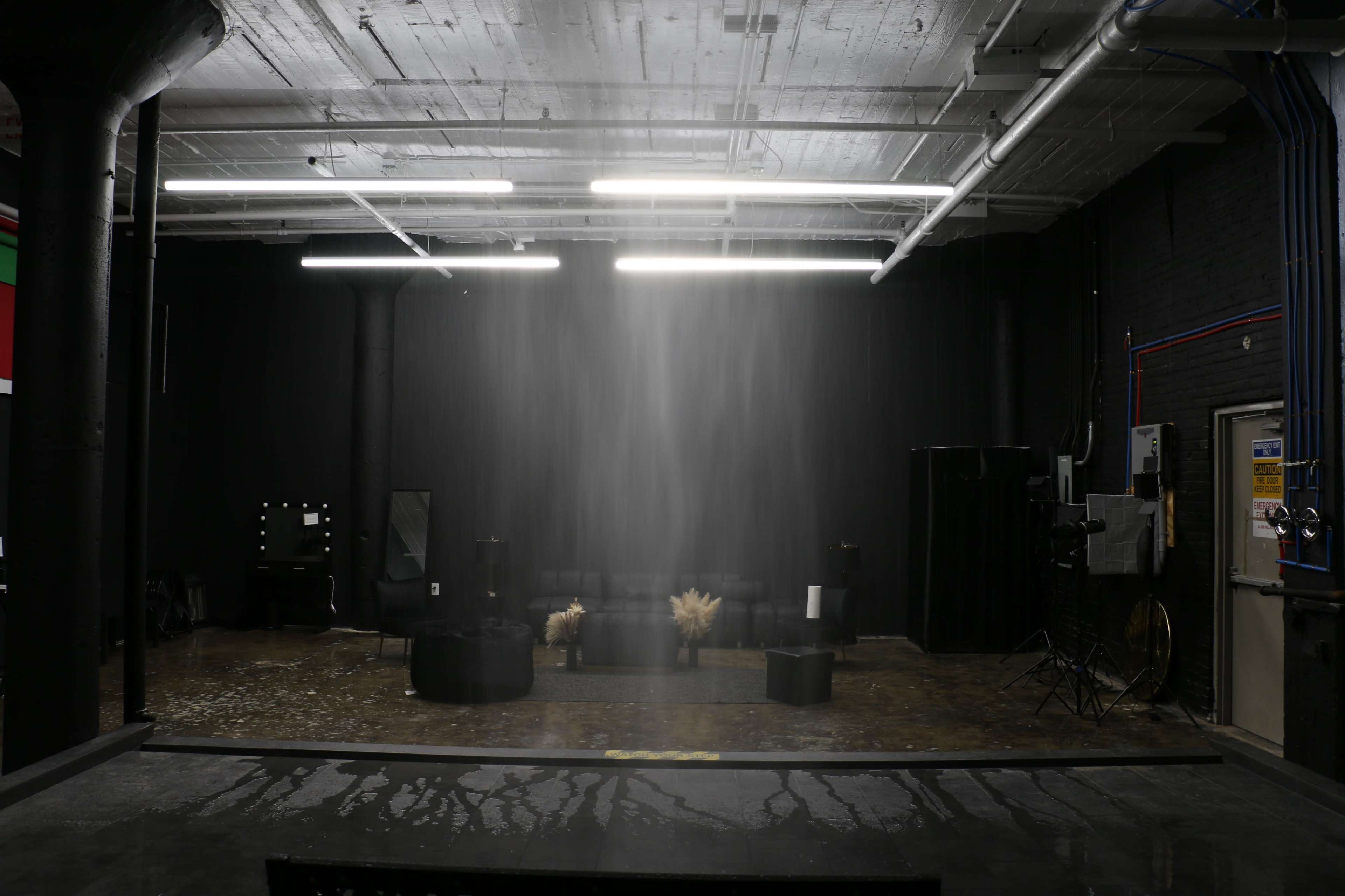 A large, dimly lit industrial space features a black backdrop with a sofa and several cylindrical tables, while water appears to cascade from the ceiling.