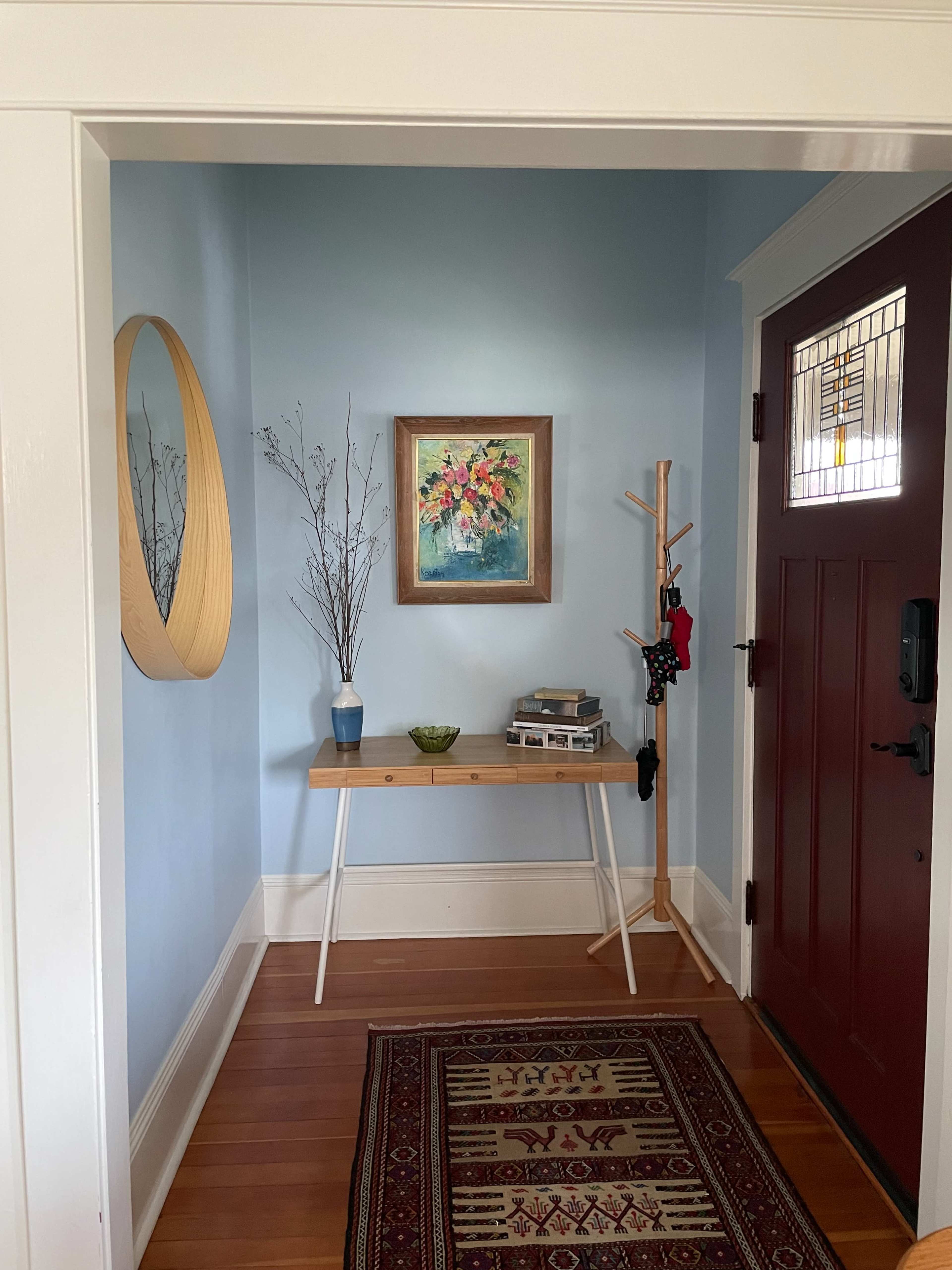 A well-lit entryway with a blue wall, a round mirror, a floral painting, a small table with a plant and books, a coat rack, and a wooden door.