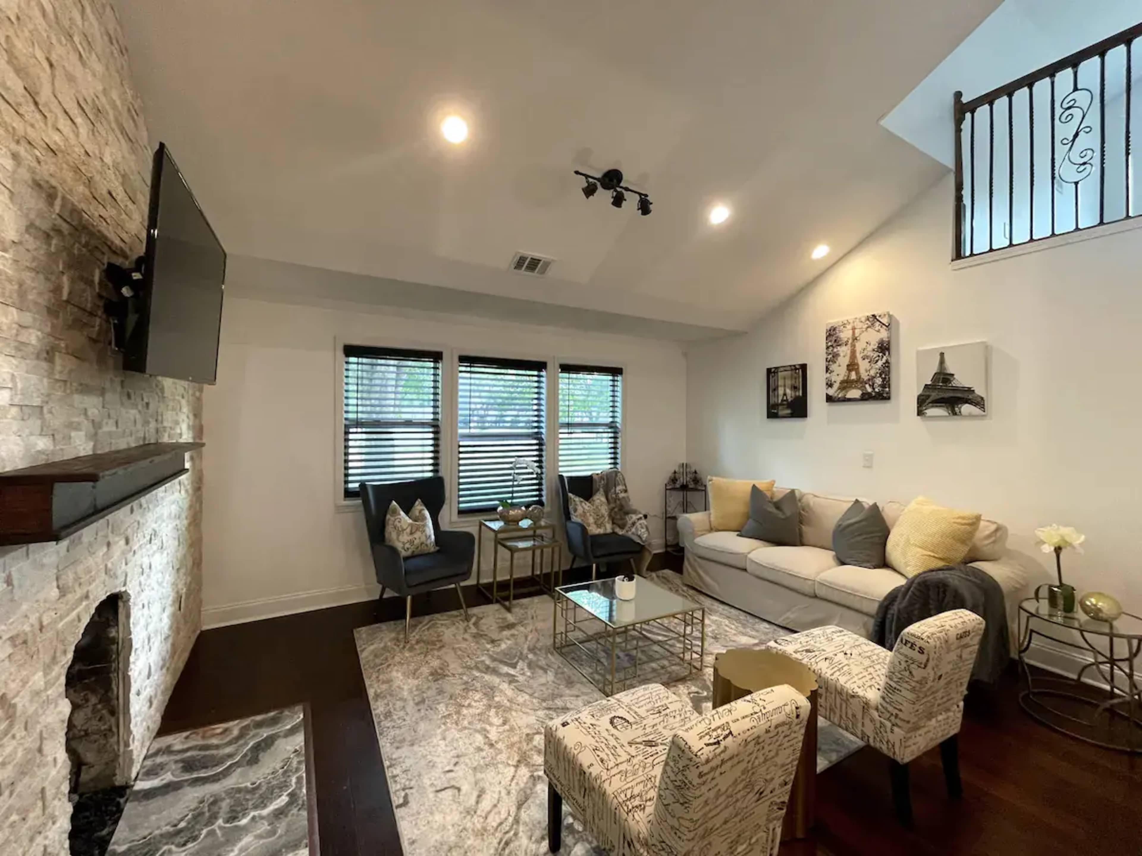 The living room features a light-colored sofa, two patterned chairs, a coffee table, and a stone fireplace, with large windows covered by black blinds.
