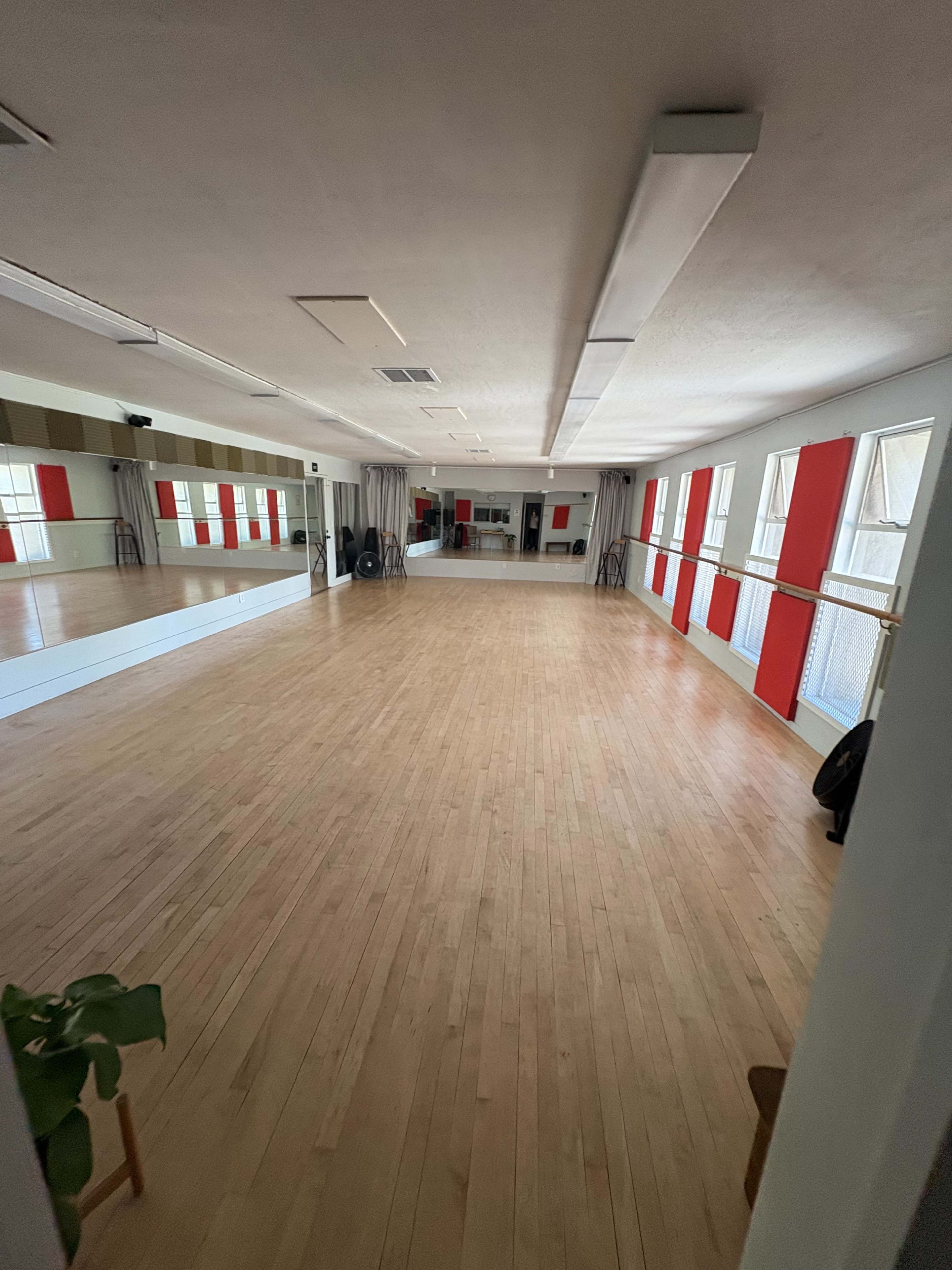 The image shows a spacious, empty dance studio with wooden flooring, large windows, and mirrored walls.
