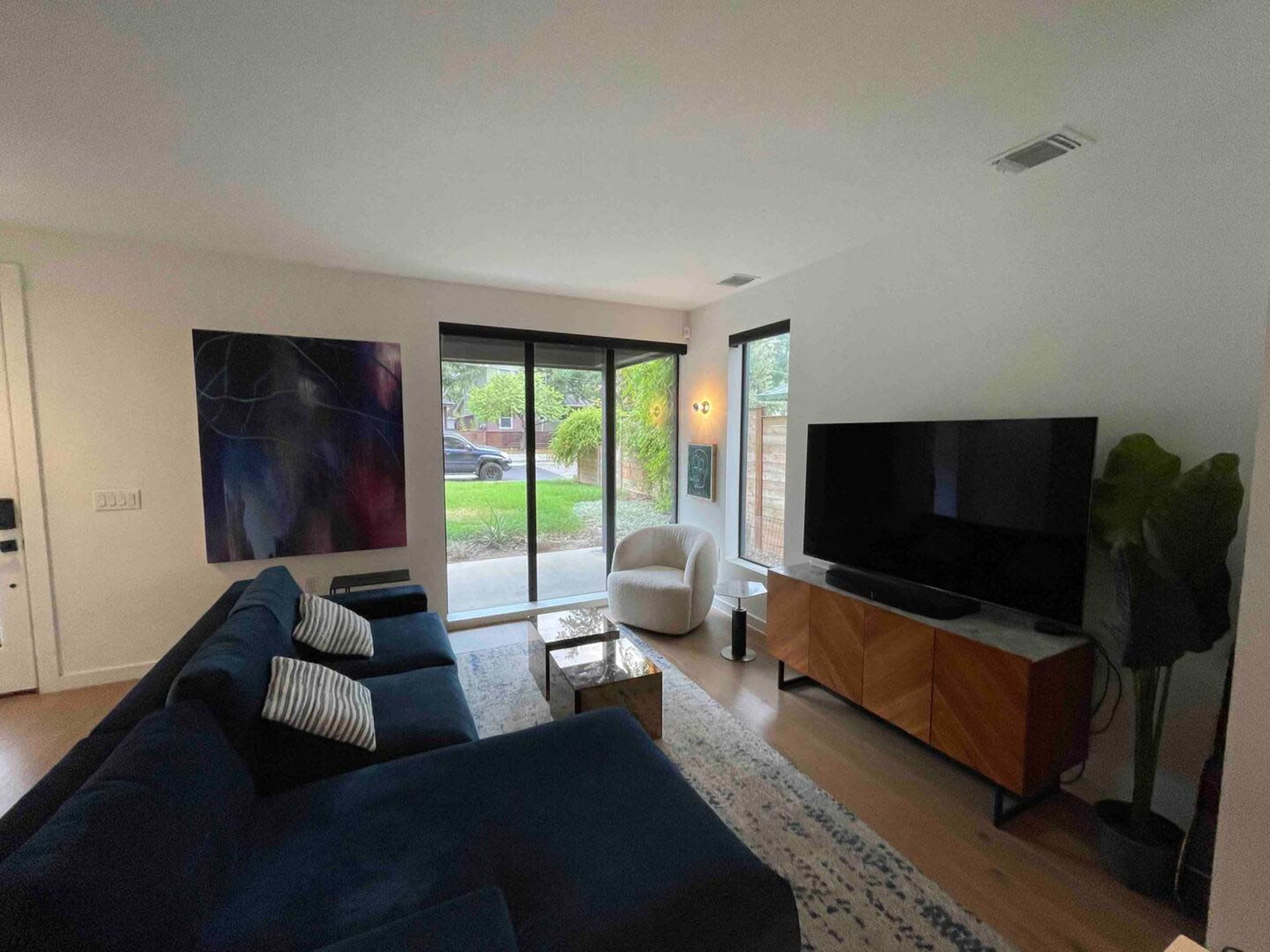 The image shows a modern living room featuring a dark blue sectional sofa, a television on a wooden cabinet, and large windows overlooking a garden.