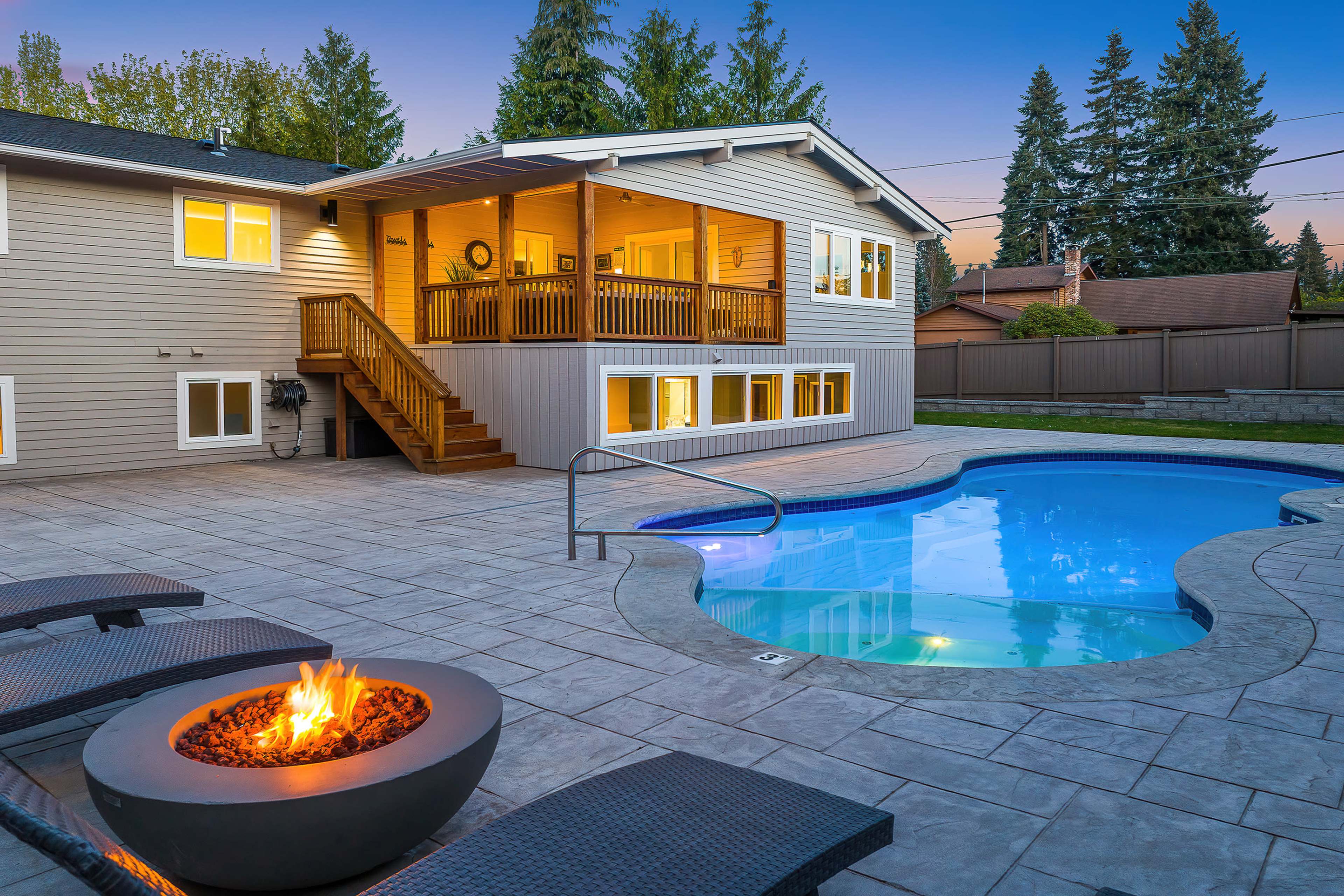 A modern home features a patio with a swimming pool, a fire pit, and a spacious deck in the background surrounded by trees.