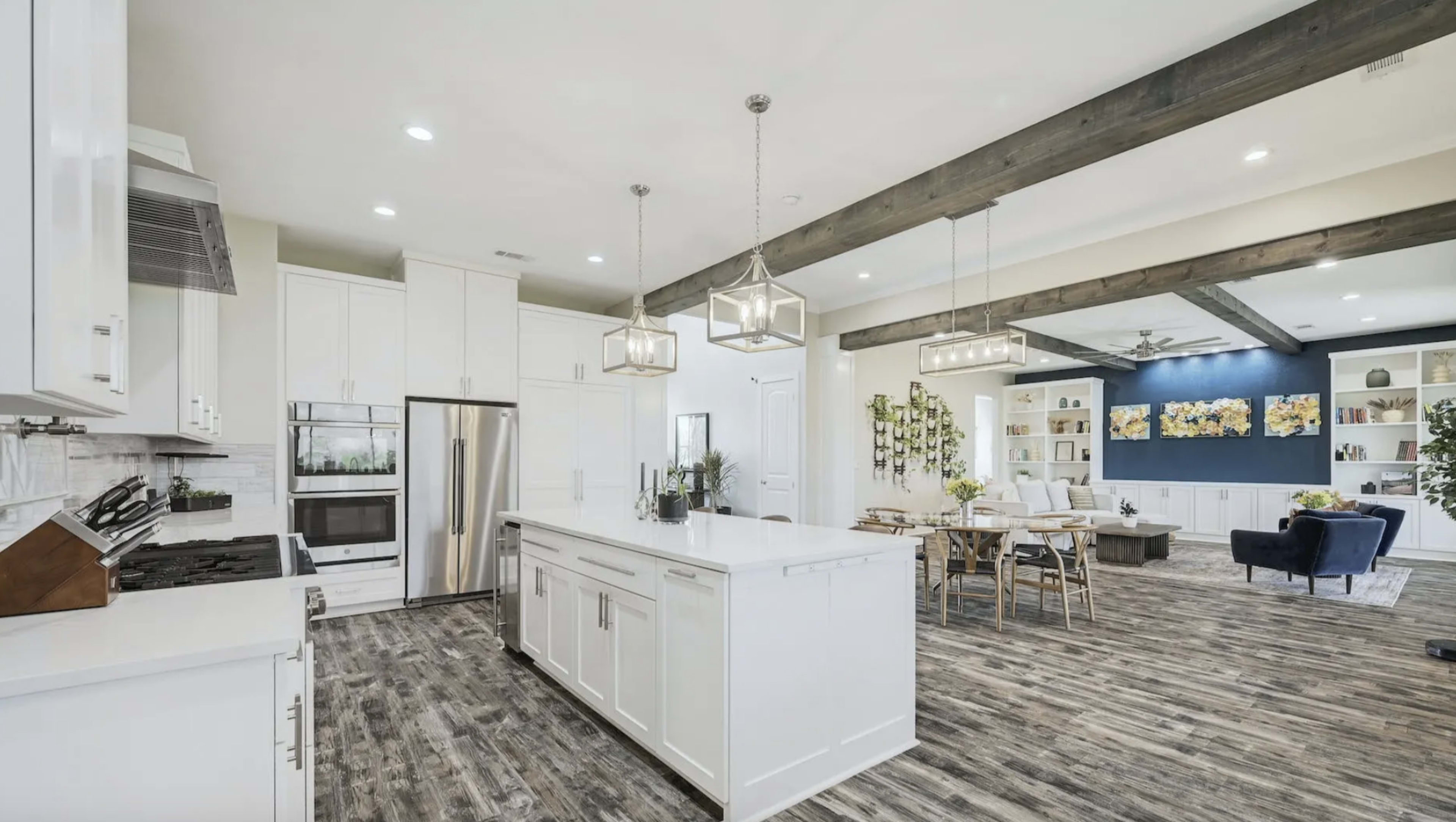 The image shows a modern kitchen with white cabinets, a large island, and hardwood flooring, connected to a dining area and living room featuring wooden beams and light-colored furniture.