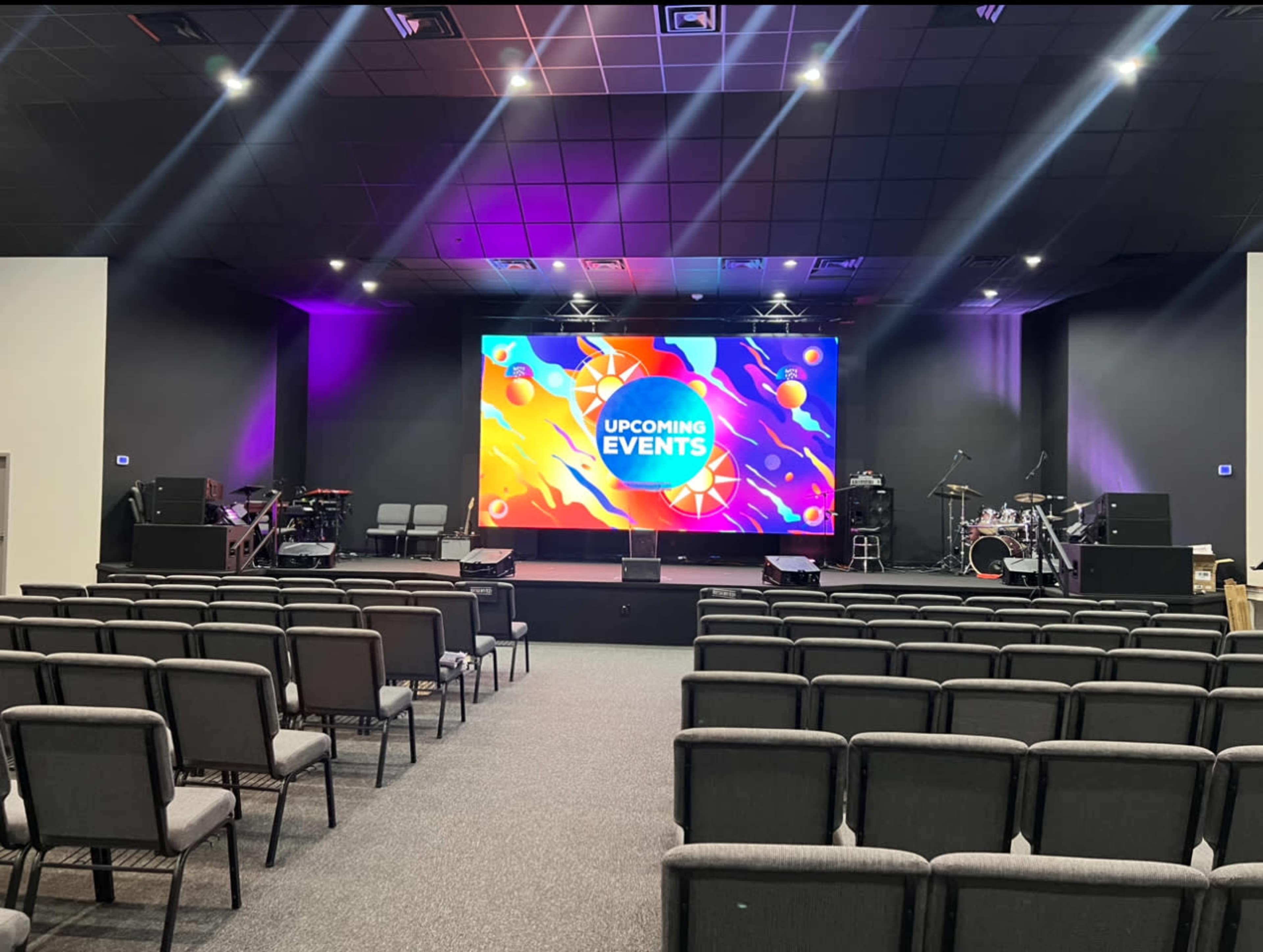 A performance space with several rows of gray chairs facing a large screen displaying "UPCOMING EVENTS," alongside musical instruments set up on stage.