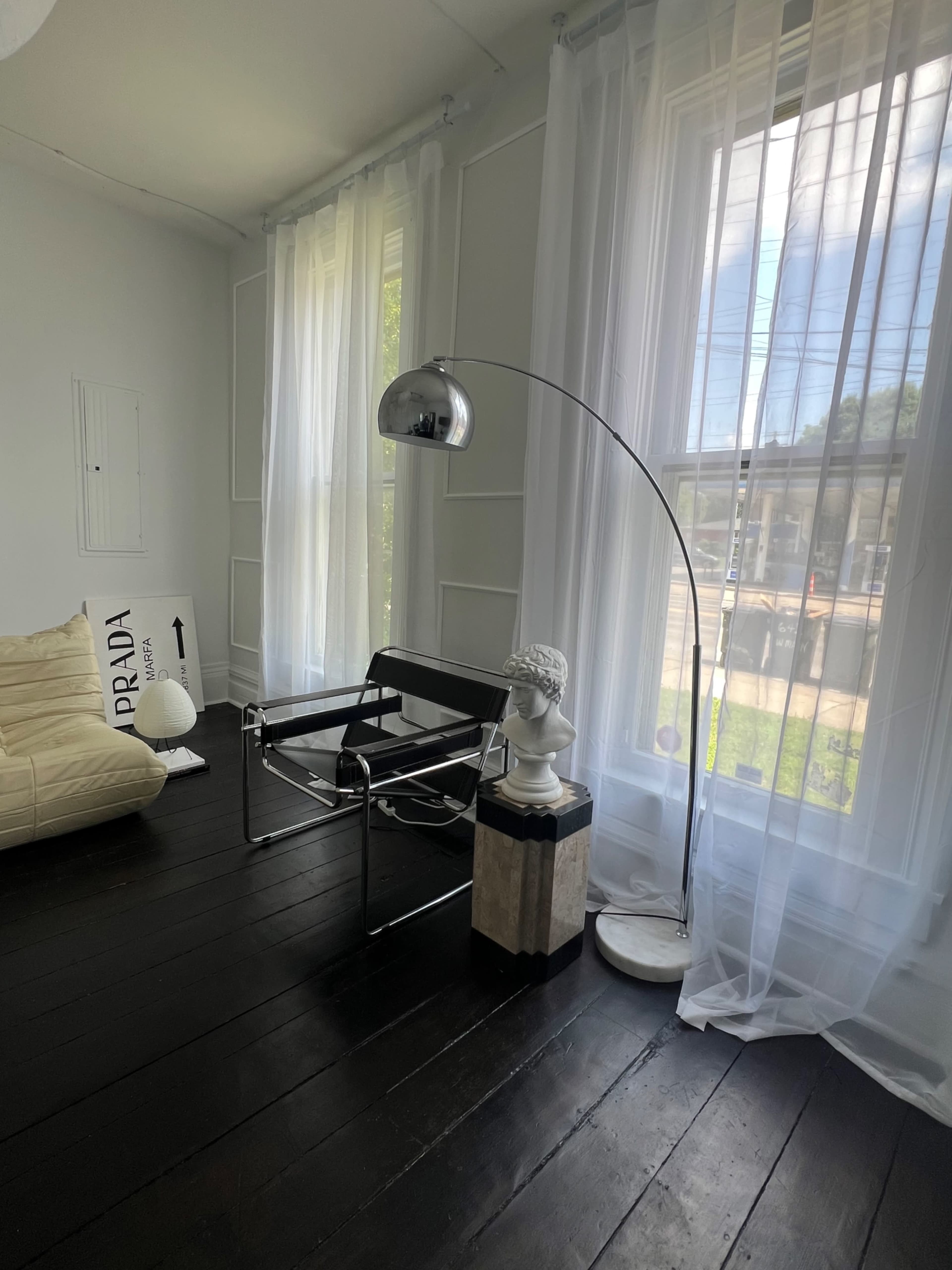 The image shows a minimalist room featuring a modern chair, a distinctive arc floor lamp, a marble pedestal with a bust, and sheer white curtains.