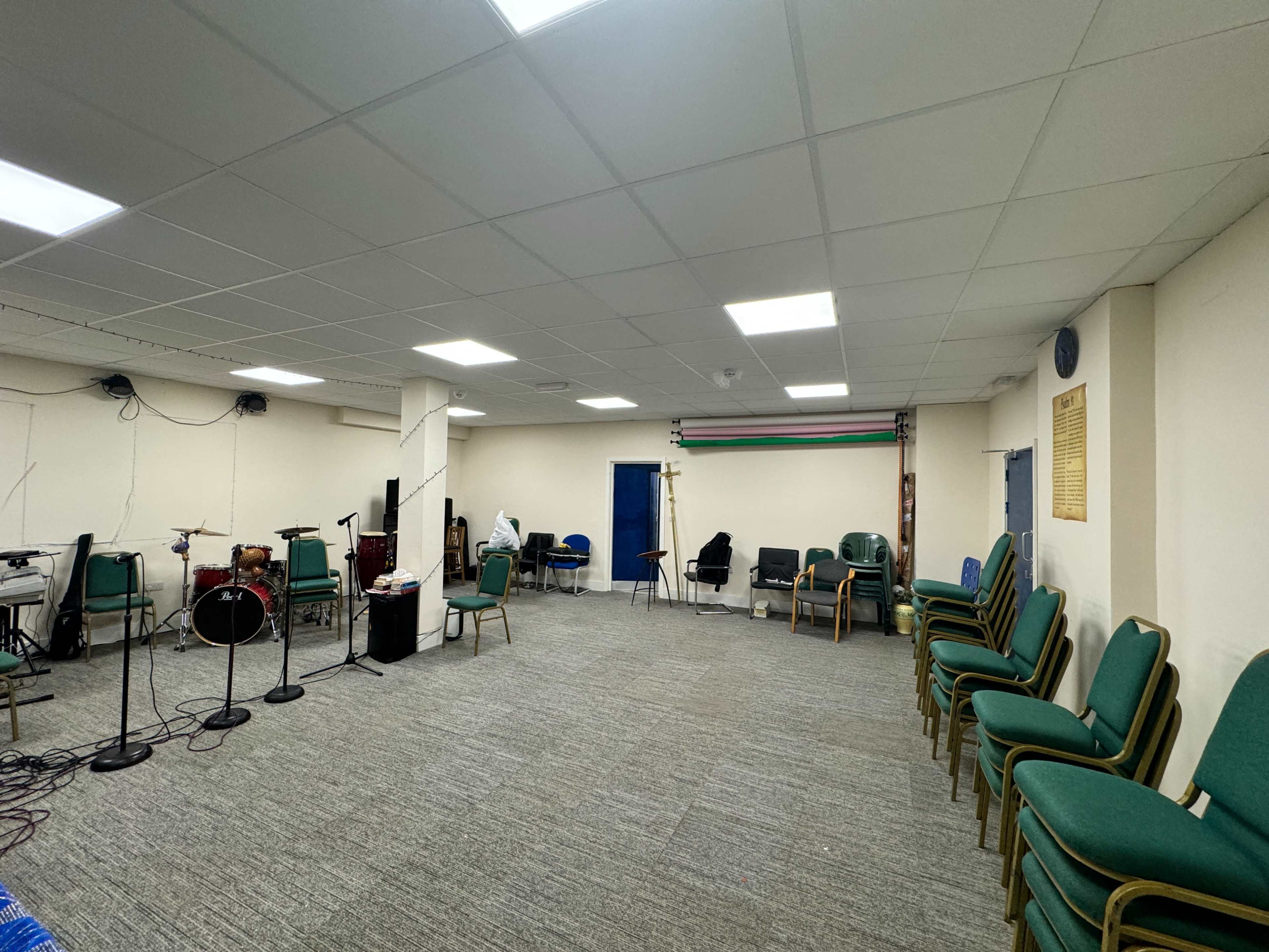 A spacious room equipped for music practice, featuring a drum set, several chairs, and a blue door leading to another area.
