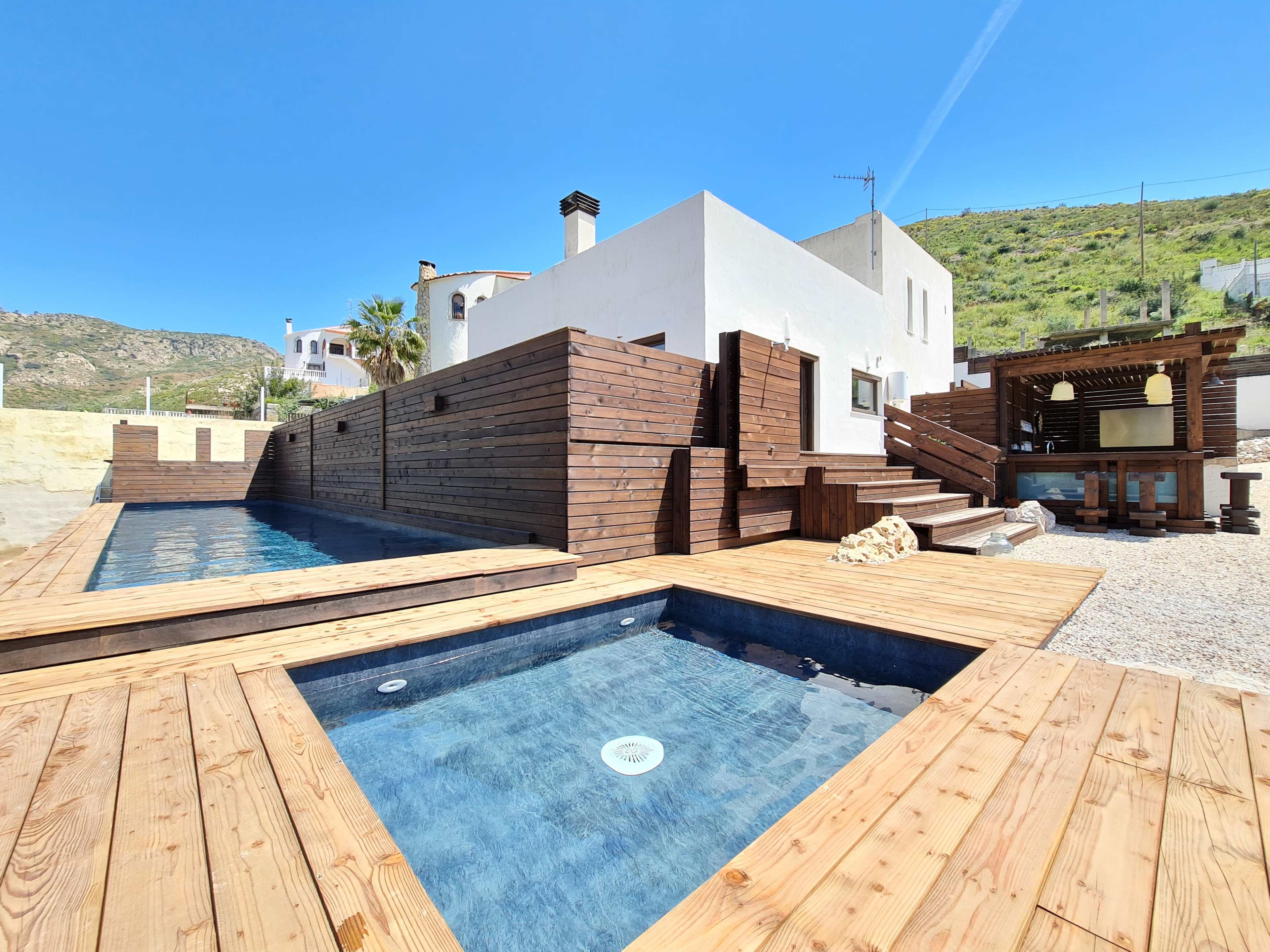 A modern house with wooden decking, featuring two small pools surrounded by a landscaped area under a clear blue sky.