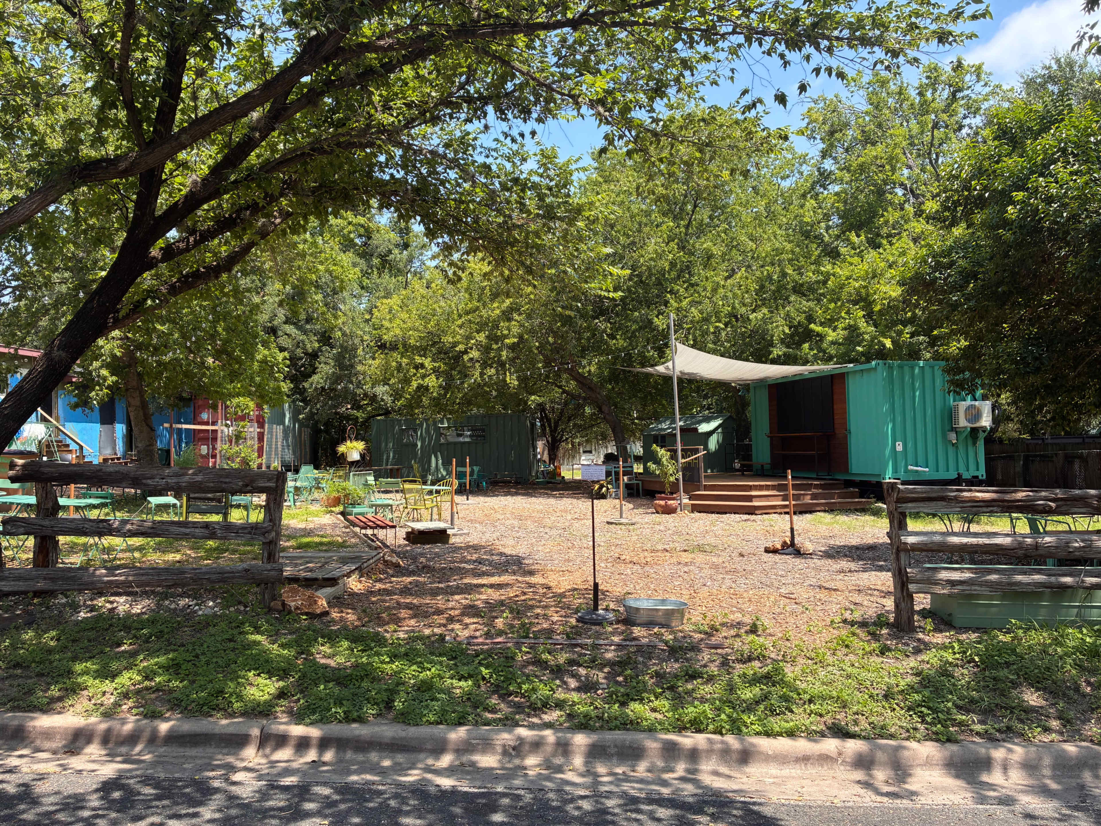 The image shows a landscape with several shipping containers, shaded by trees, set in a gravel area with benches and outdoor seating.
