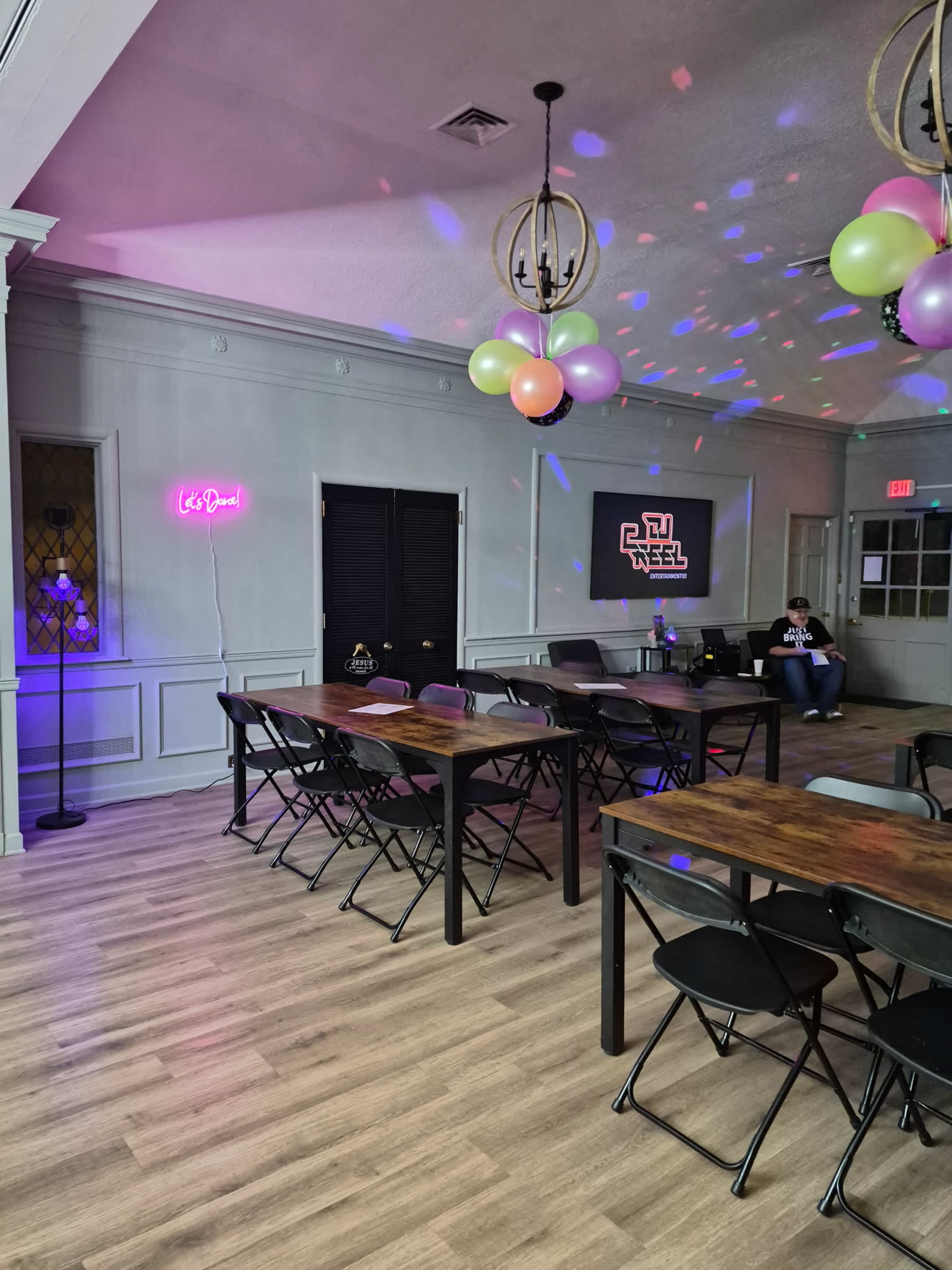 A brightly lit room features several tables and chairs, colorful balloons hanging from the ceiling, and a neon sign that reads "Let's Dance!" on one wall.