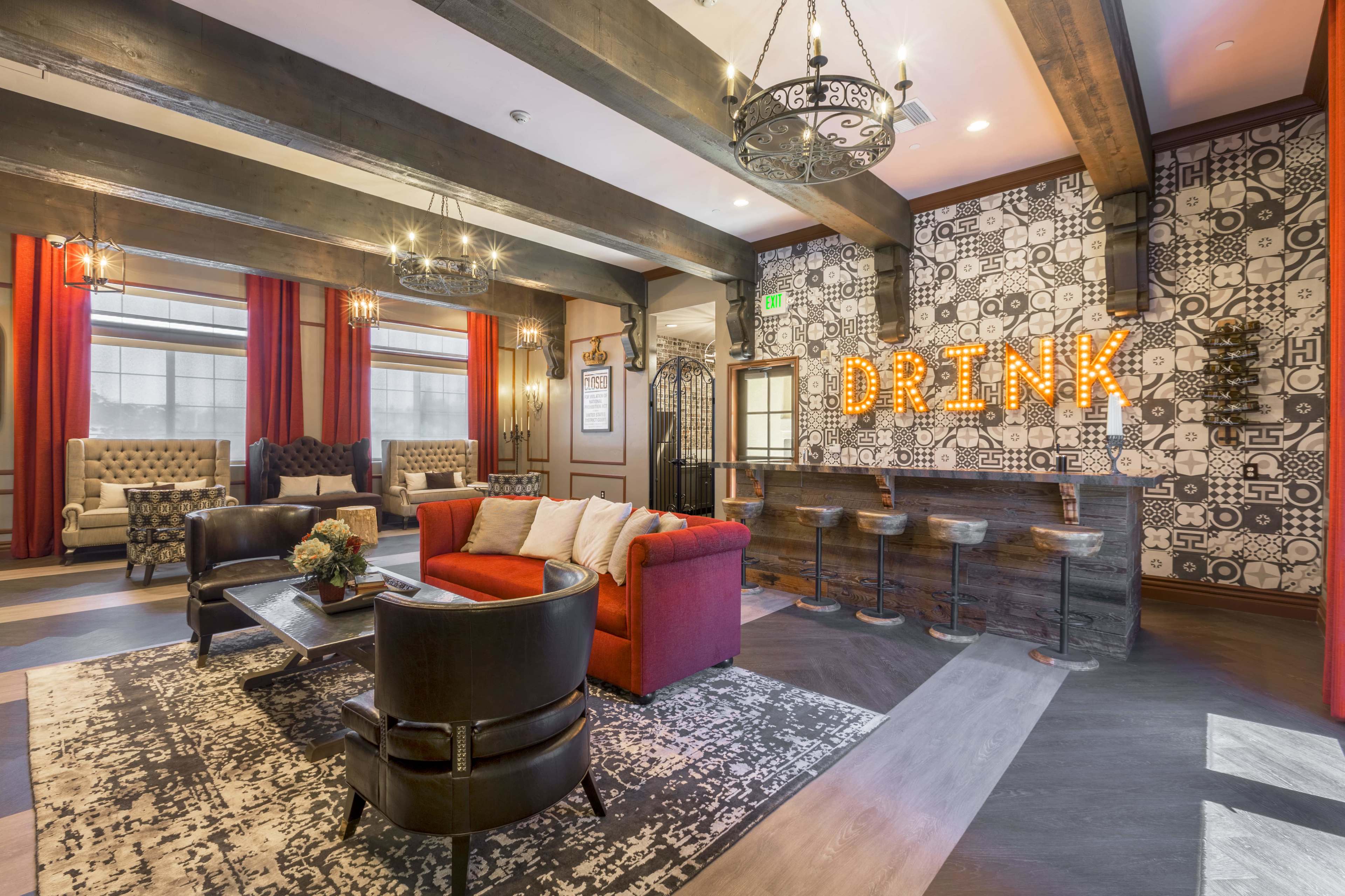 A modern lounge area features a red sofa, black chairs, and a bar with illuminated "DRINK" signage against a patterned wall.