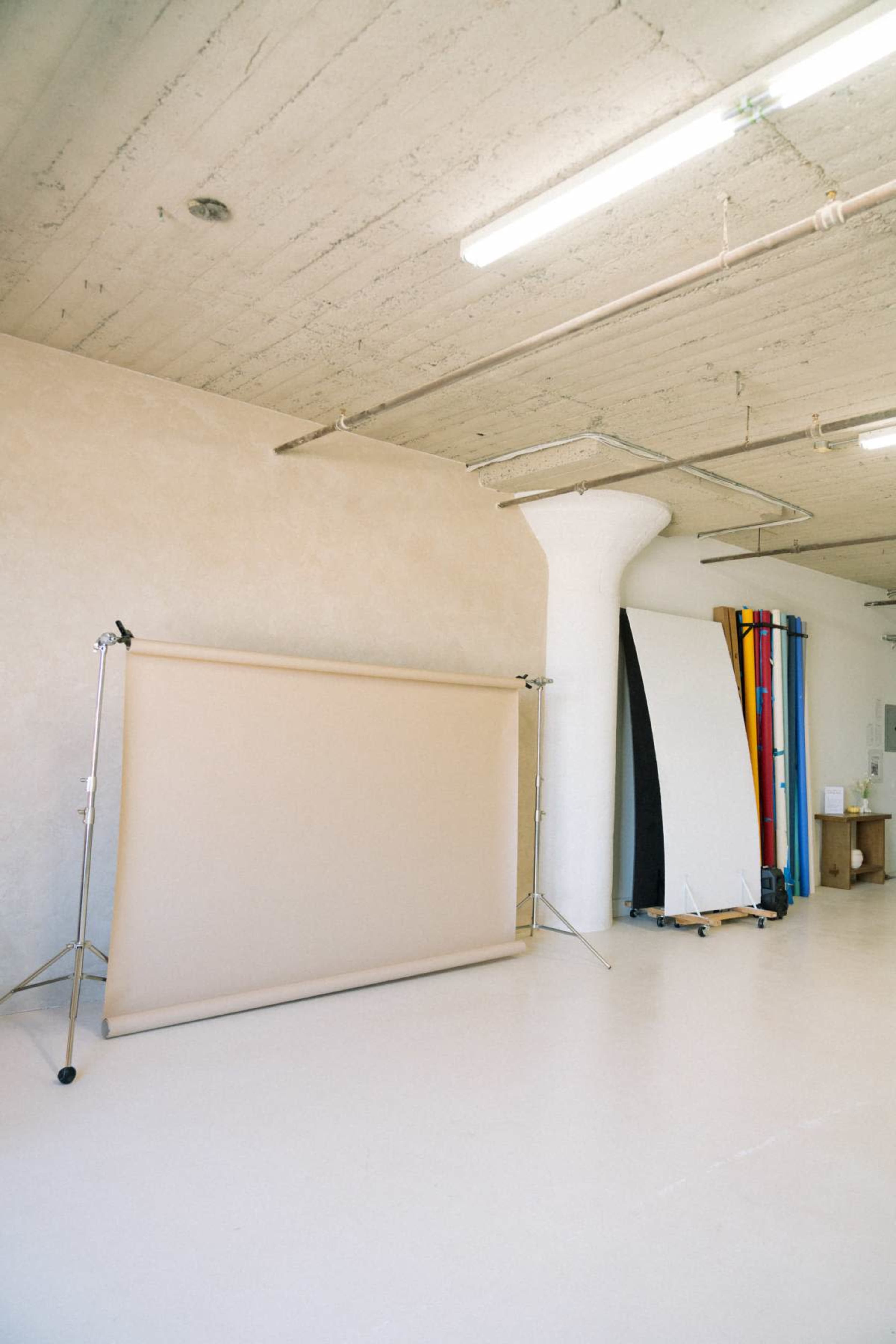 The image shows a photography studio featuring a backdrop stand with a beige backdrop and several color backdrops leaning against a wall.