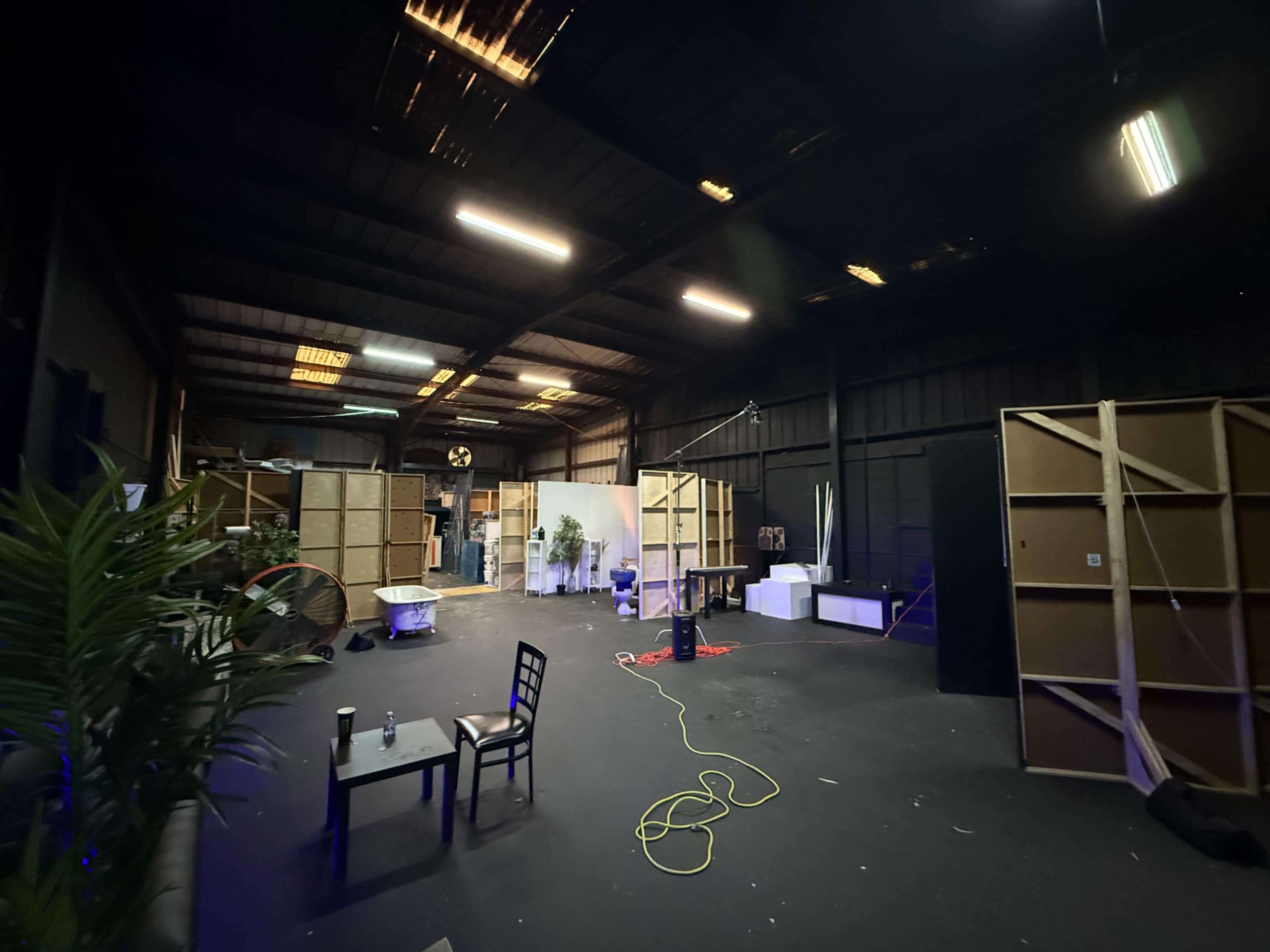 The image shows an unfinished industrial space with wooden partitions, a table, chairs, and various lighting setups scattered throughout.