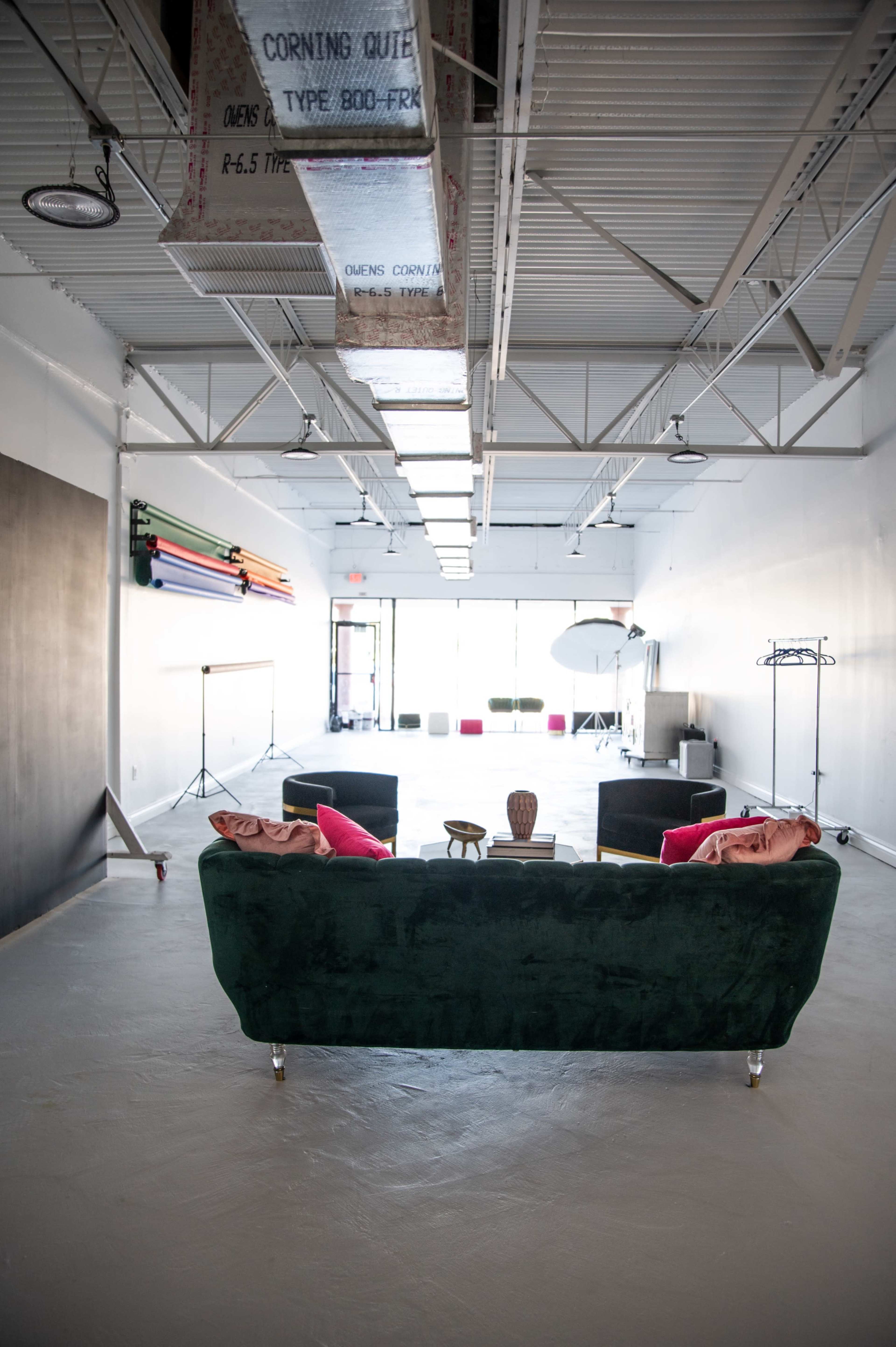 The image shows an empty, well-lit creative studio space featuring a green couch, colorful backdrop supports along the wall, and various lighting equipment.