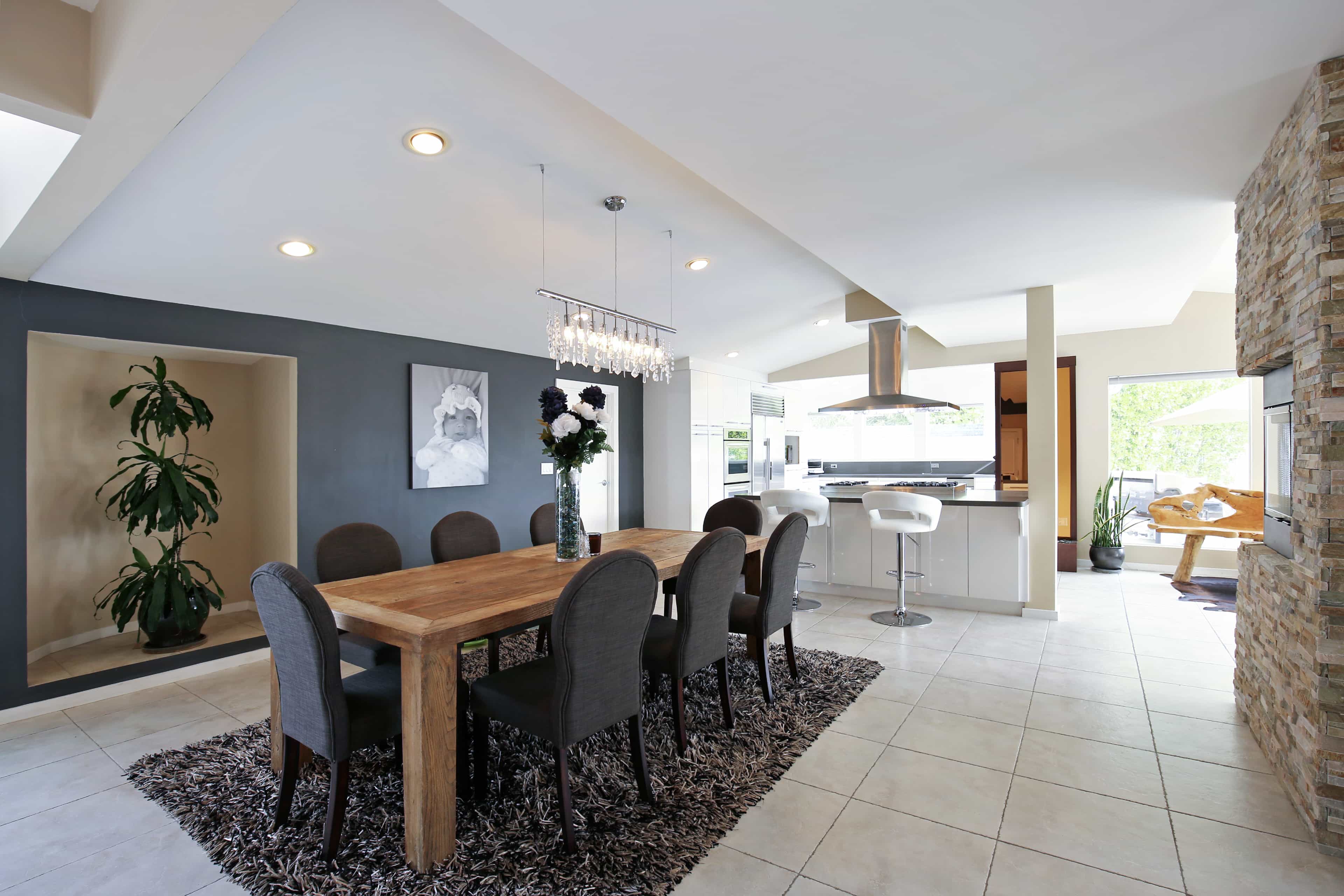 The image shows a modern dining area with a wooden table and six chairs, adjacent to a kitchen with a stone accent wall and a large window.