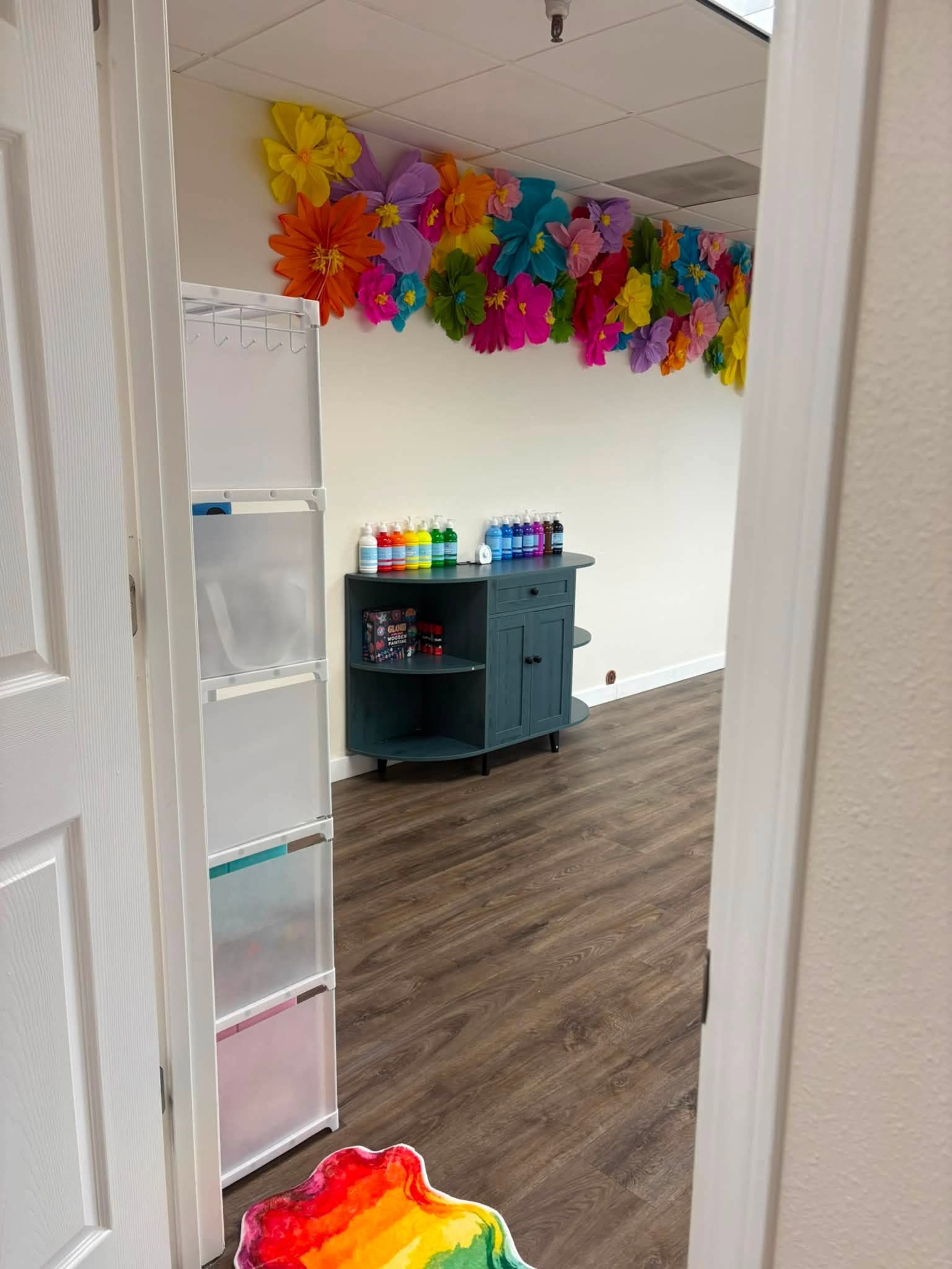 The image shows the interior of a room with a colorful floral wall decoration, a dark furniture piece displaying bottles of paint, and a rainbow-patterned mat at the entrance.