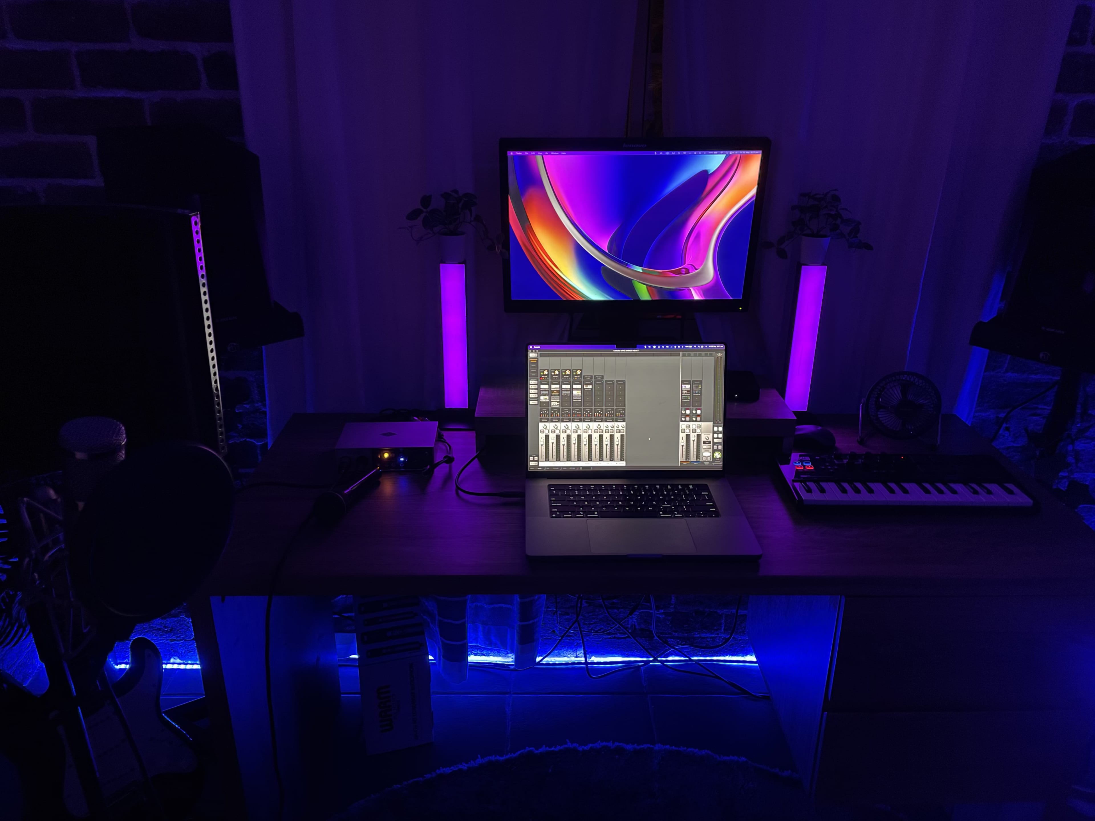 A music production setup features a laptop on a wooden desk, flanked by studio monitors and a colorful computer display, all illuminated with blue and purple lighting.