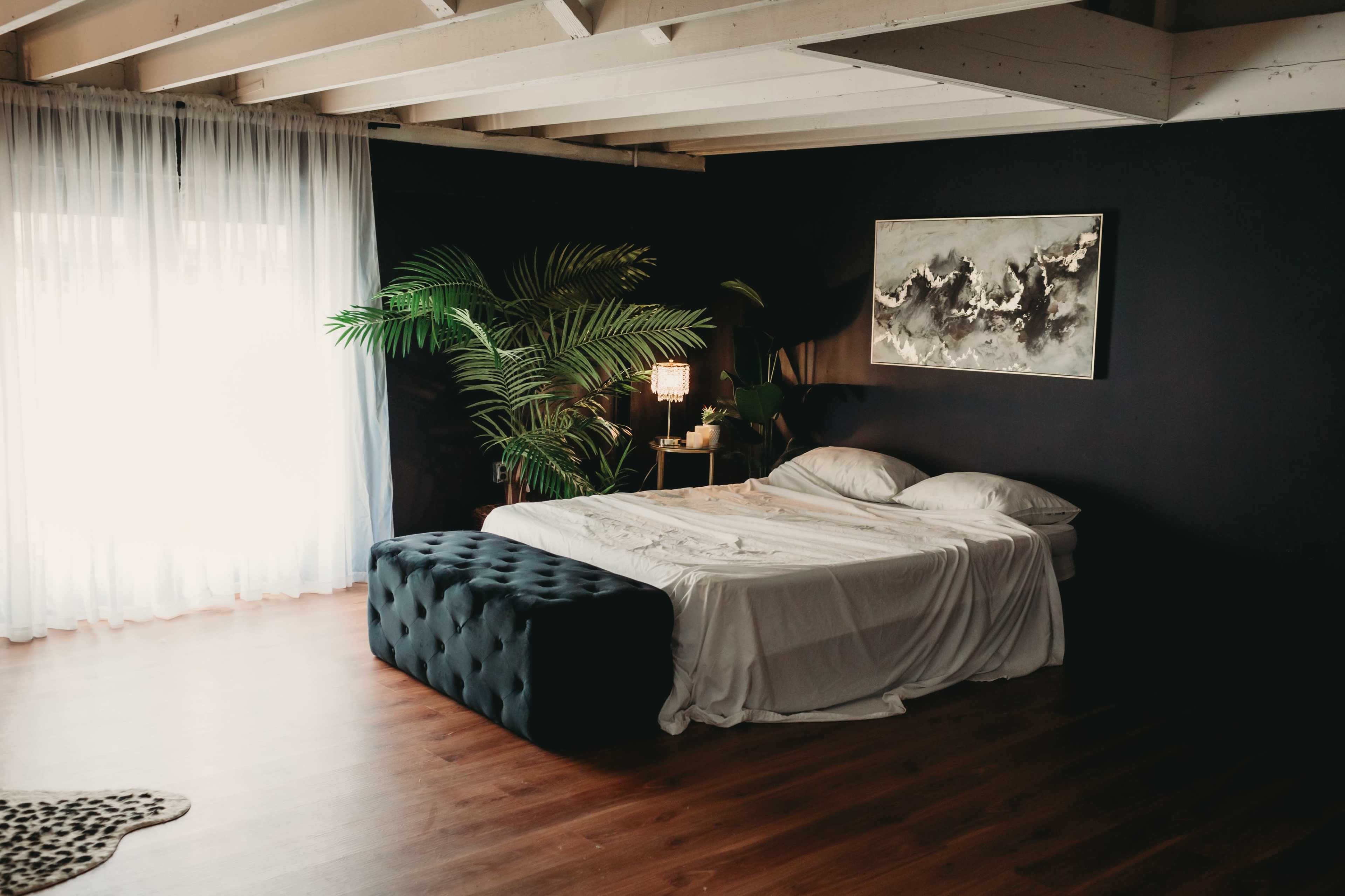 A neatly made bed is positioned near a large window with sheer curtains, accompanied by a stylish bench and a potted plant in a modern room.