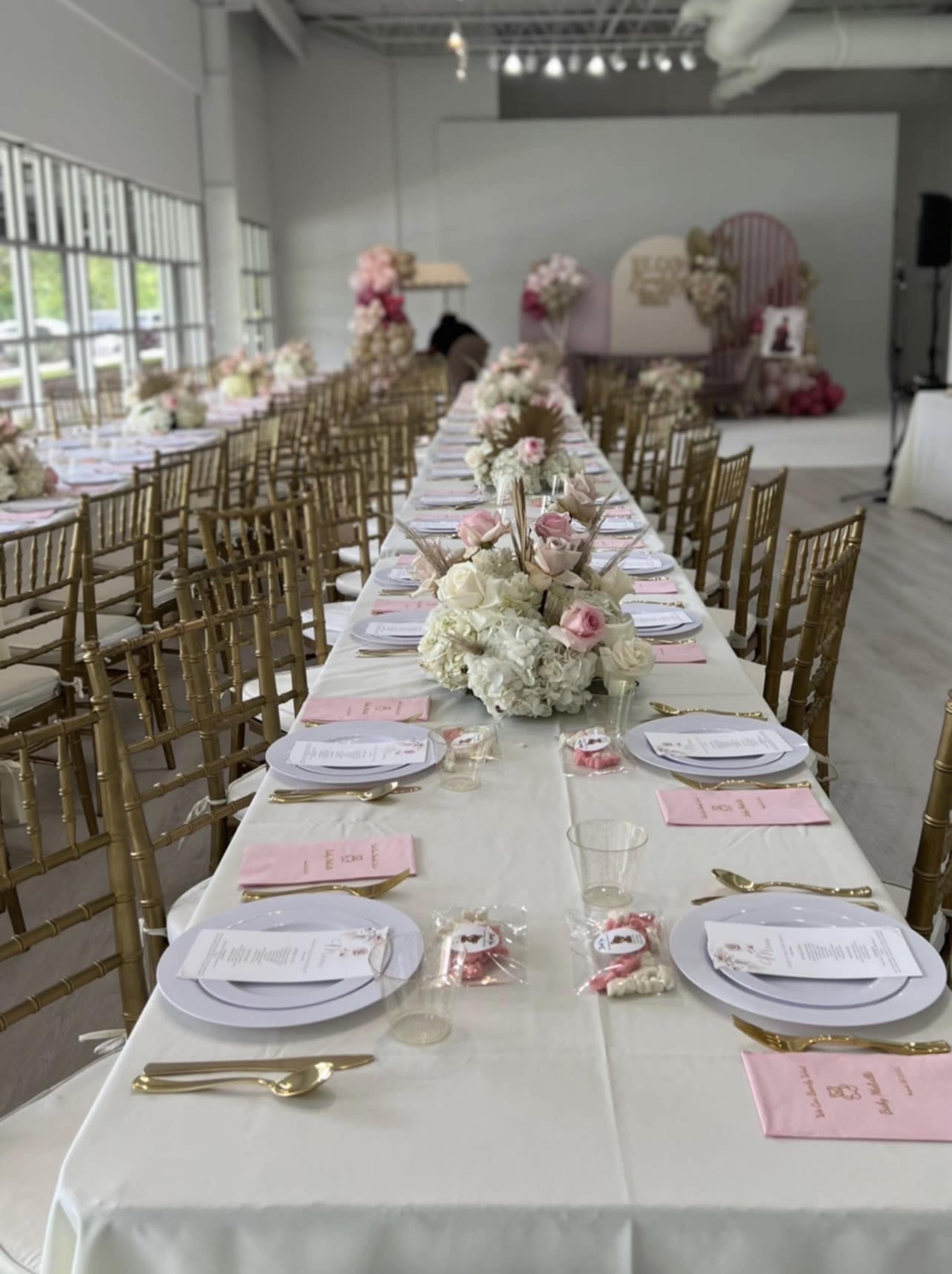 A long wedding reception table is set with white tablecloths, gold chairs, floral centerpieces, and neatly arranged plates and menus.