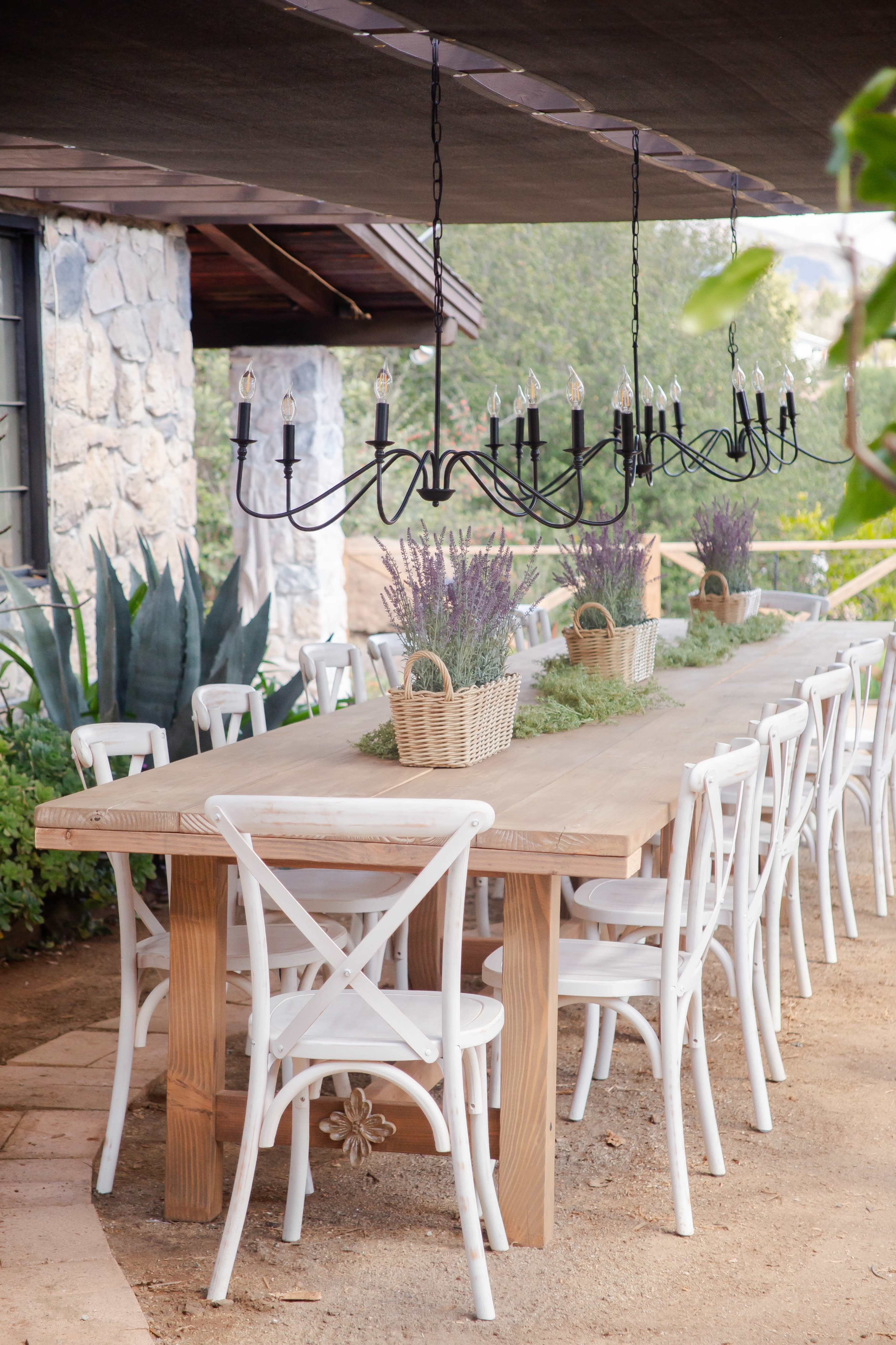 A long wooden dining table surrounded by white chairs is set under a covered patio, decorated with hanging lights and potted lavender.