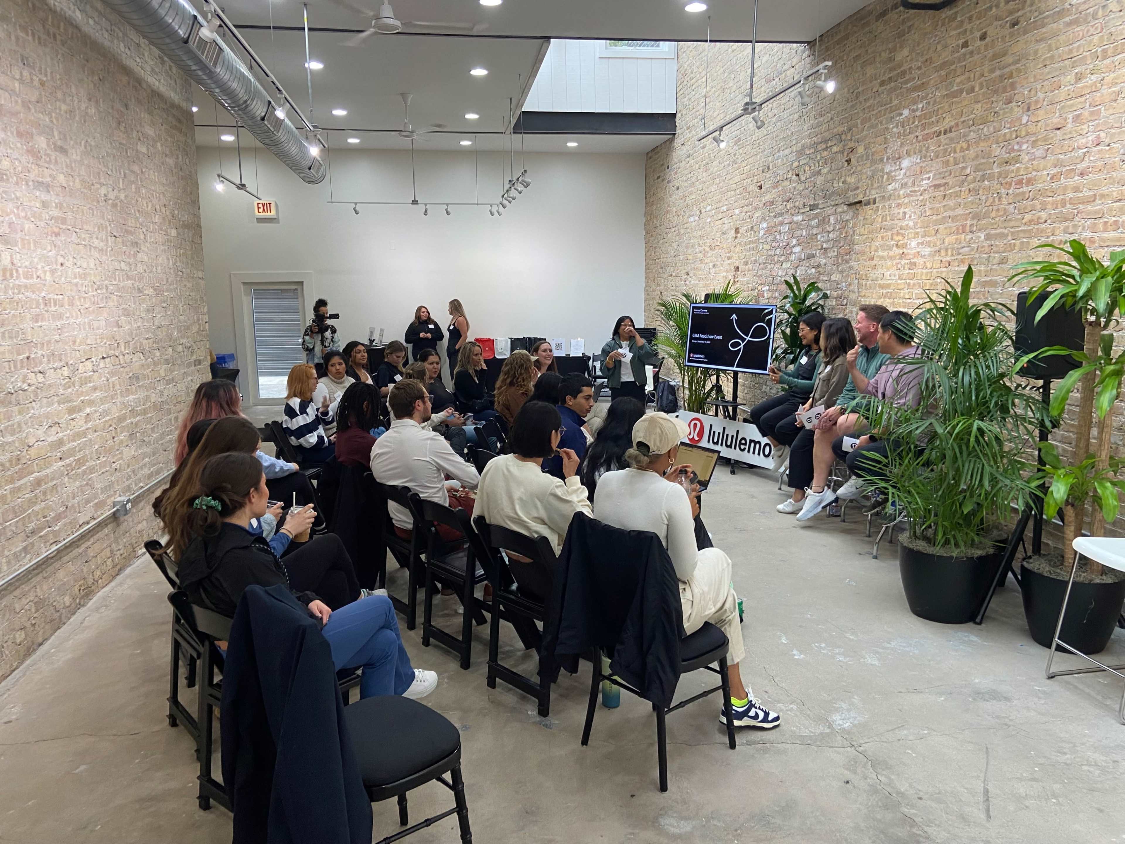 A group of people sits in chairs facing a panel discussion in a modern studio with brick walls and potted plants.