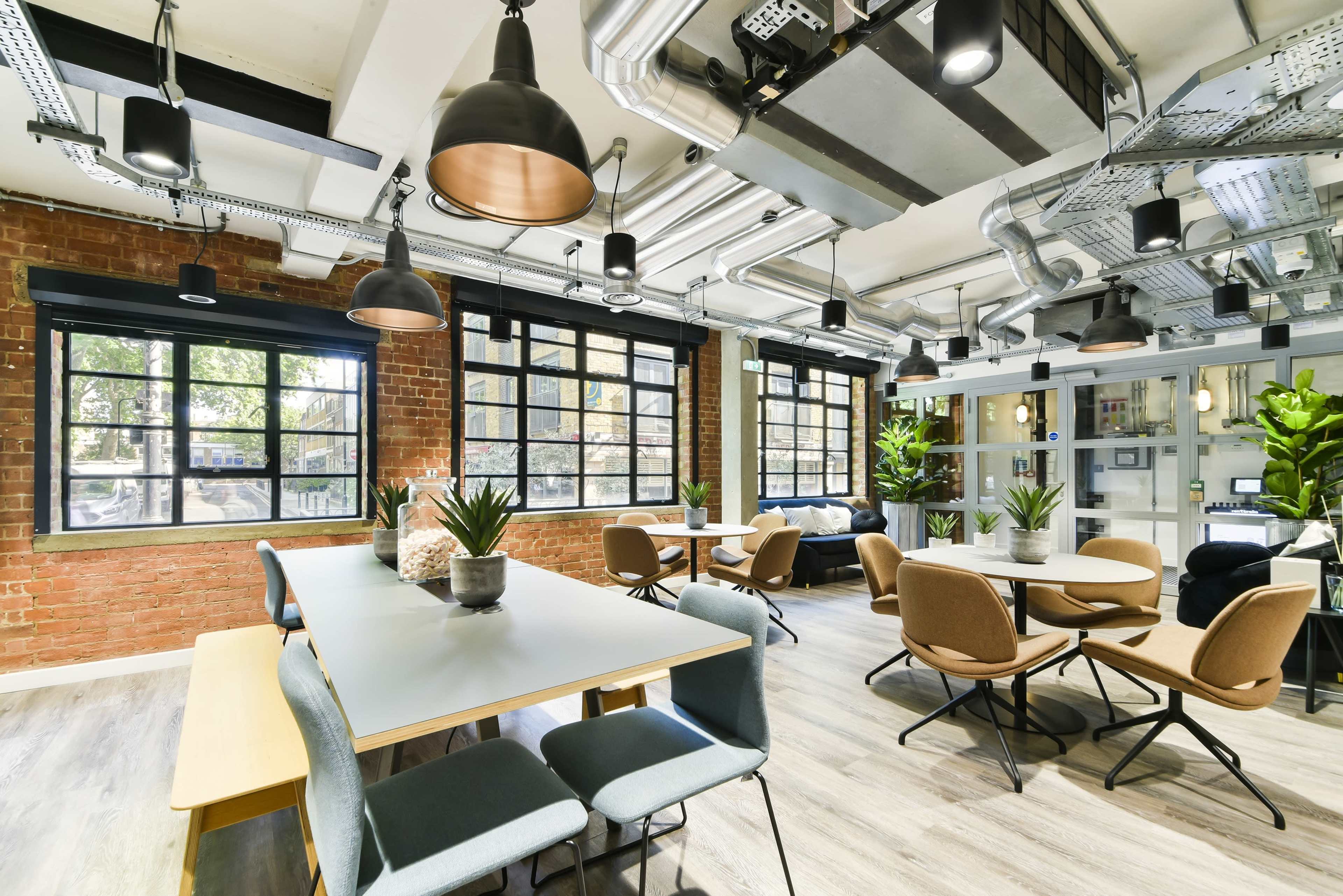 A modern workspace with large windows, industrial-style lighting, and a mix of tables and chairs arranged around potted plants.