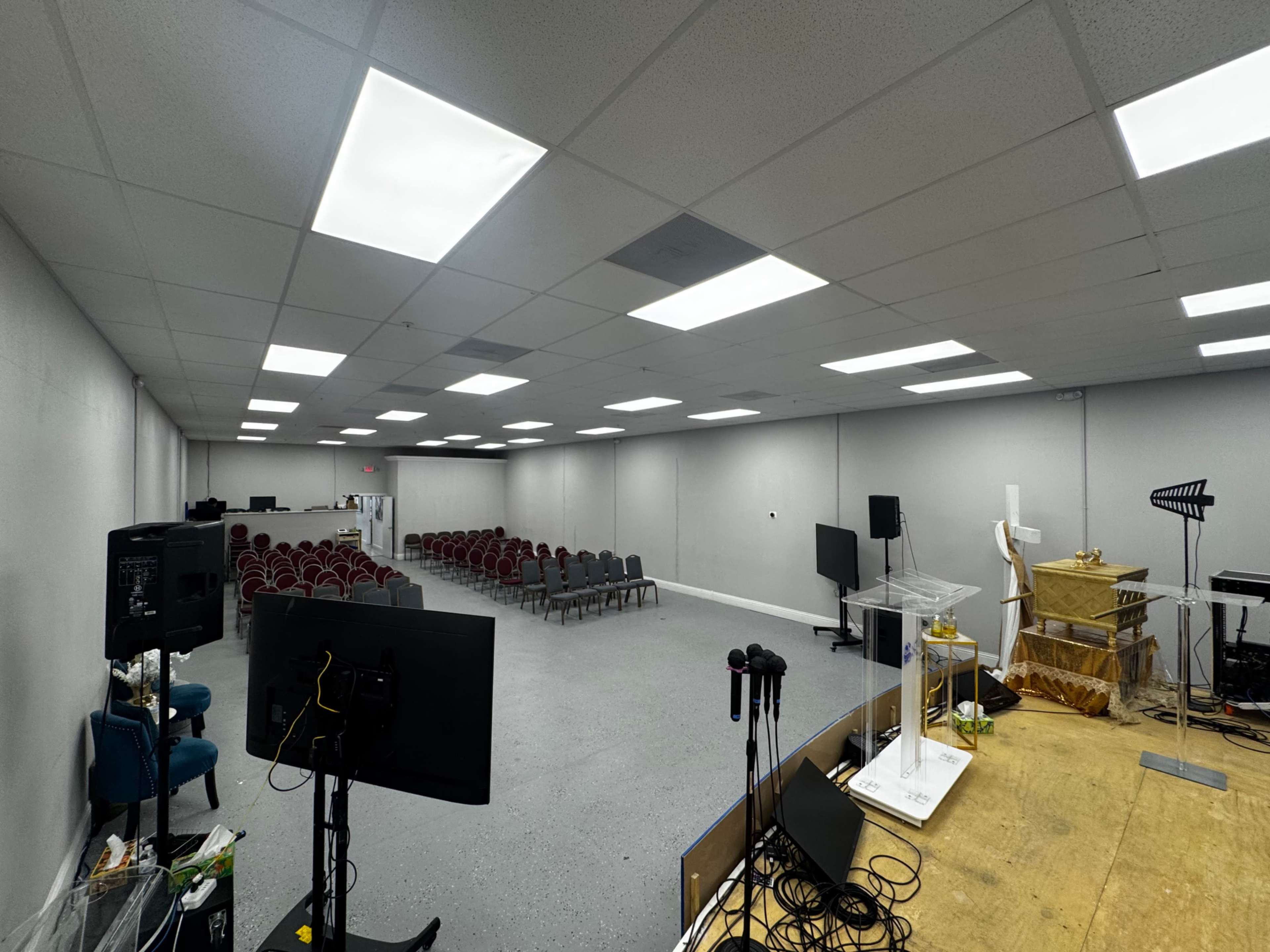 The image shows a spacious room with rows of chairs arranged for an audience, a stage with a podium, and various audio-visual equipment set up.