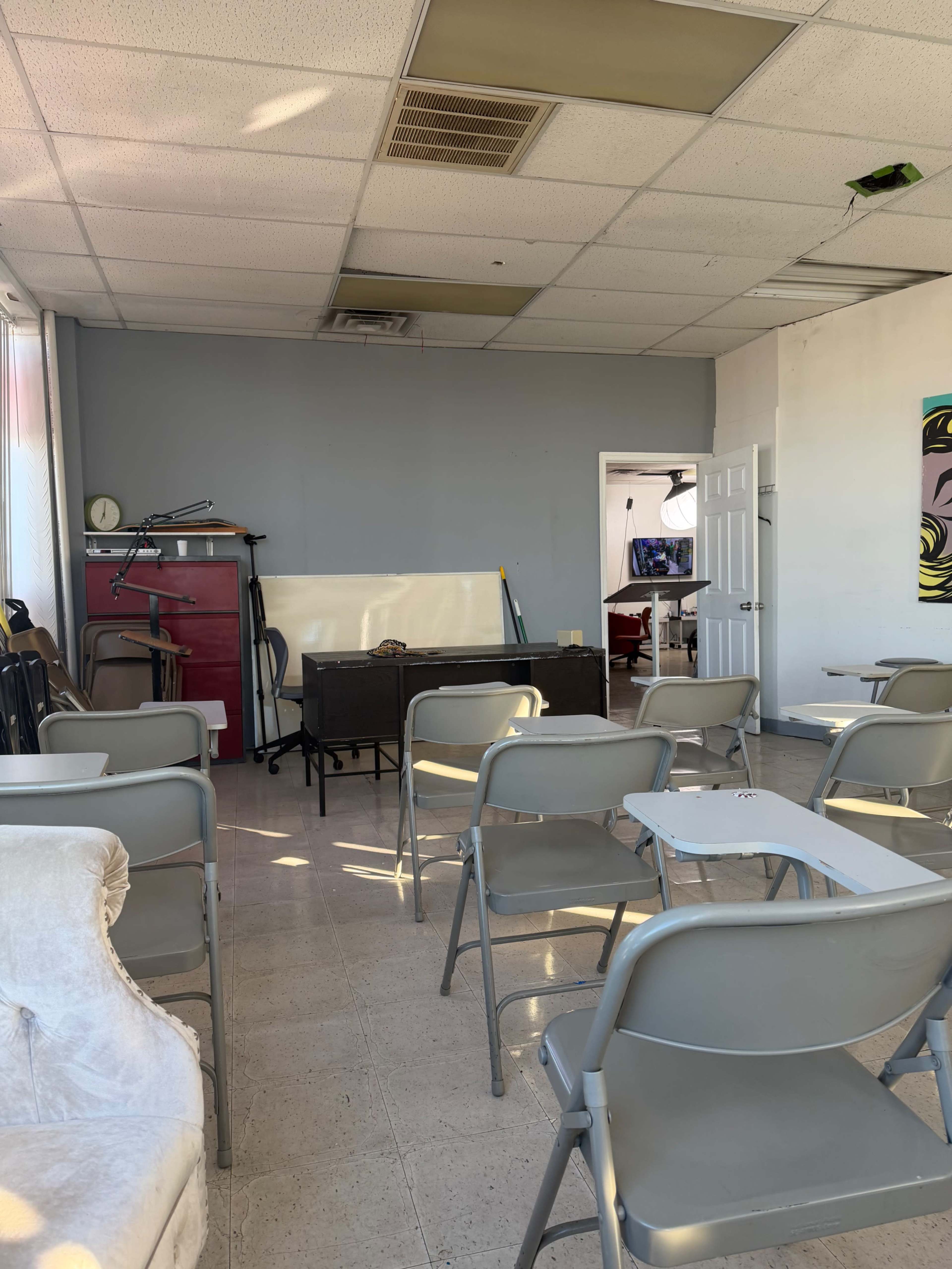 The image shows a sparsely furnished room with several grey chairs arranged in rows, a wooden desk, and a whiteboard against the wall.