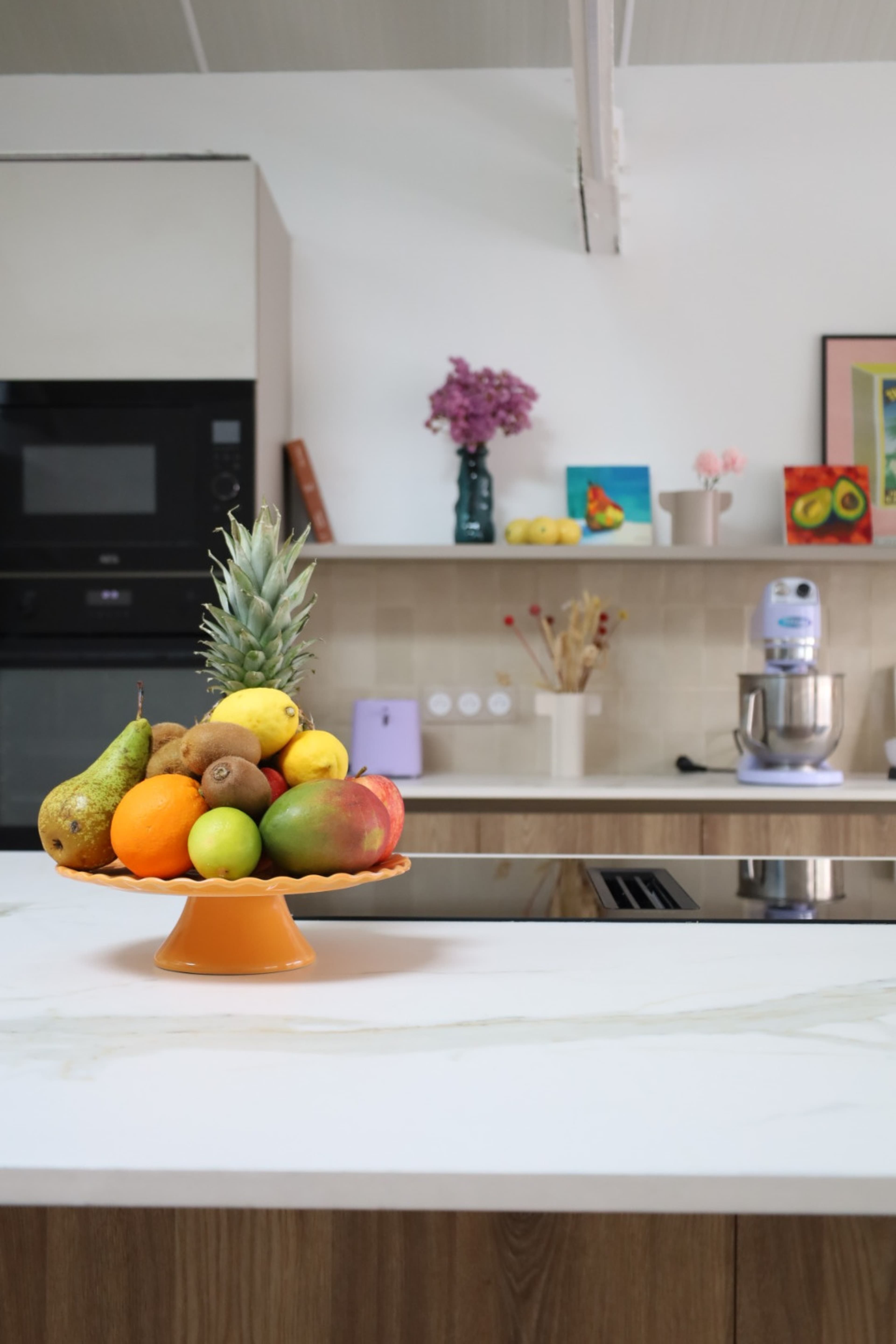 A bowl of assorted fruits, including apples, pears, and a pineapple, rests on a countertop in a modern kitchen with white cabinets and a flower arrangement in the background.
