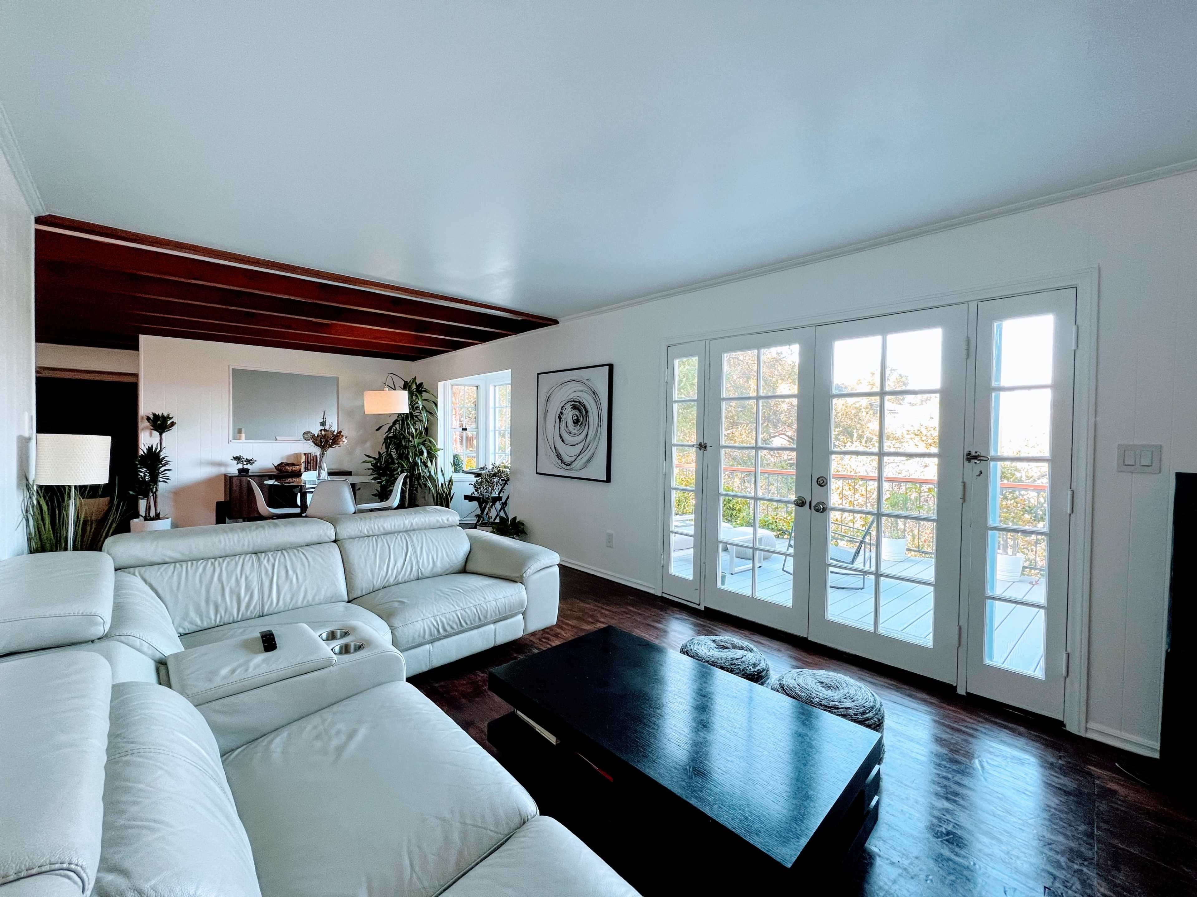 A modern living room features a white leather sectional sofa, a black coffee table, and large glass doors leading to a sunlit balcony.