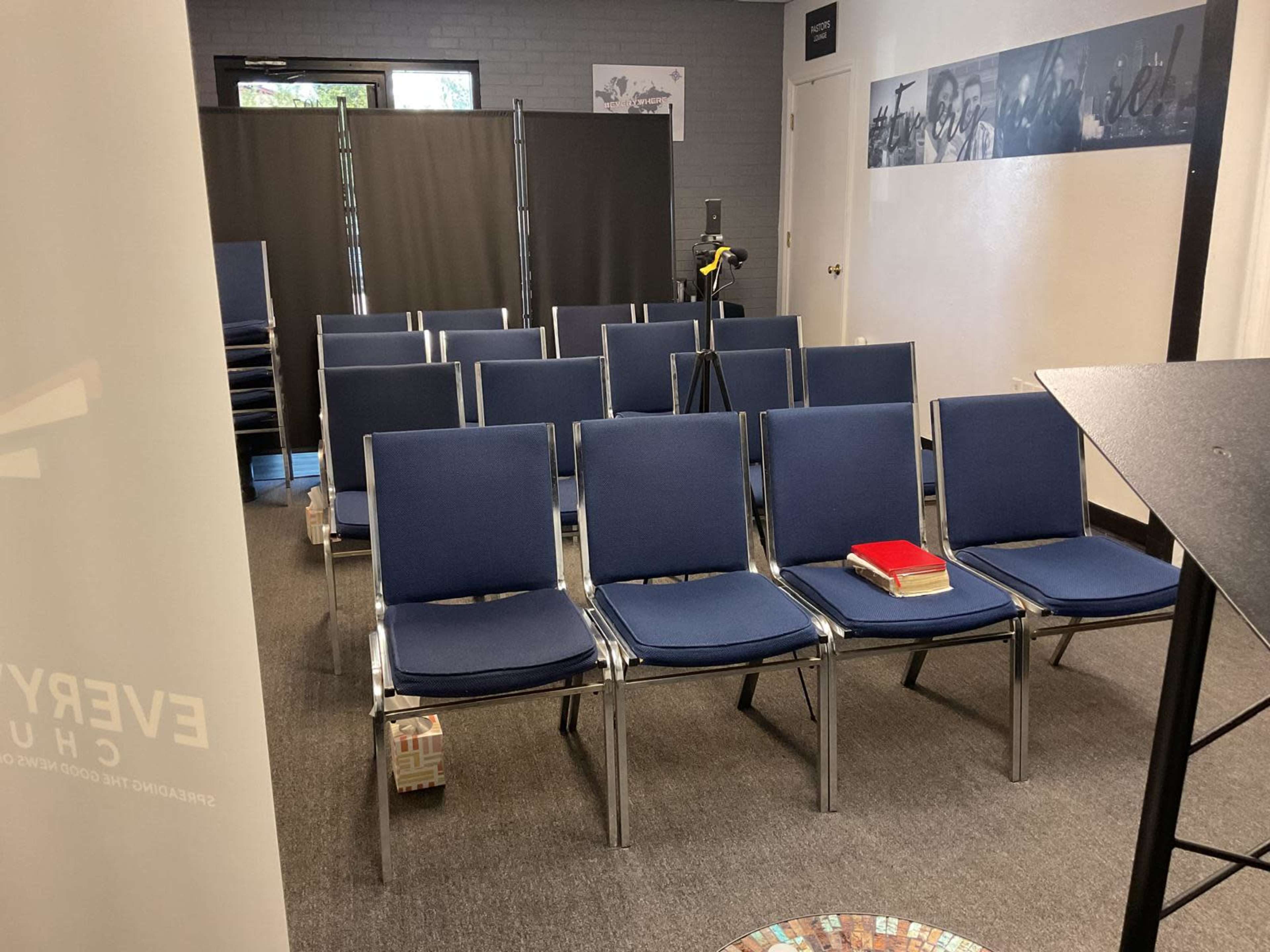 The image shows a sparsely arranged room with several rows of blue chairs facing a speaker's podium, with a few items placed on a chair.