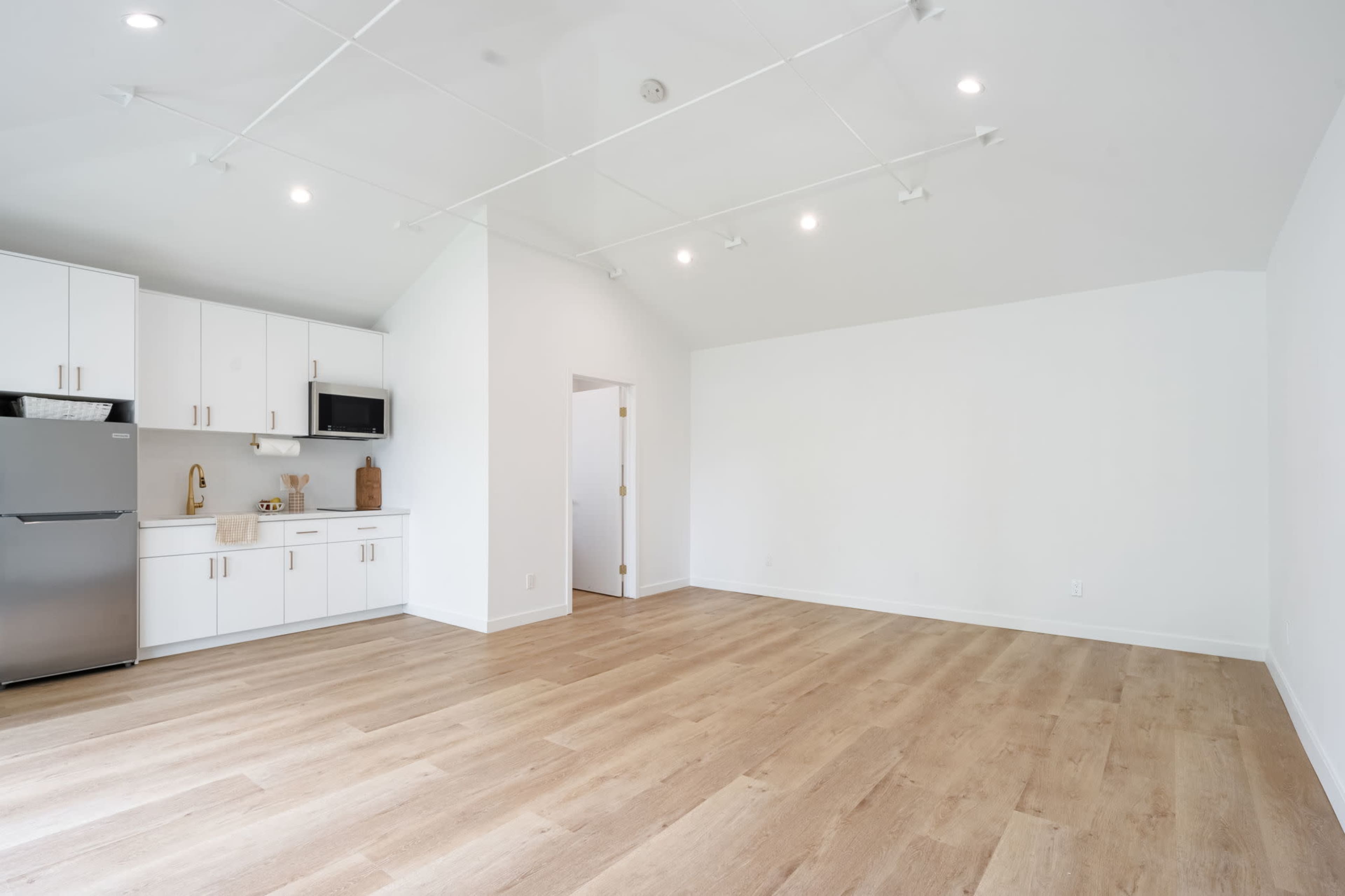A spacious, empty room features light wooden flooring, white walls, and a kitchen area with modern appliances.