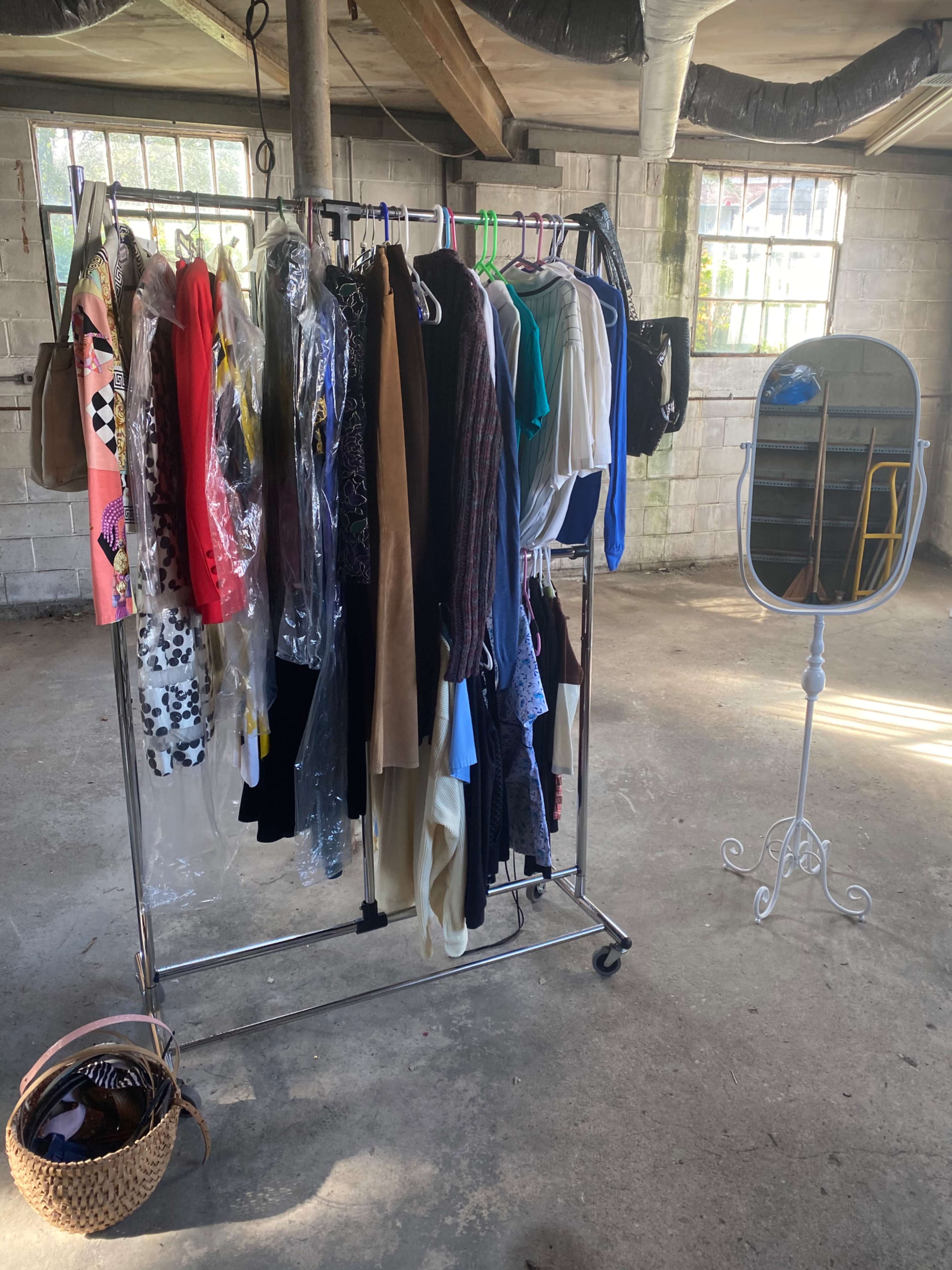 A clothing rack filled with various garments is positioned in front of a standing mirror in a sparsely furnished room.