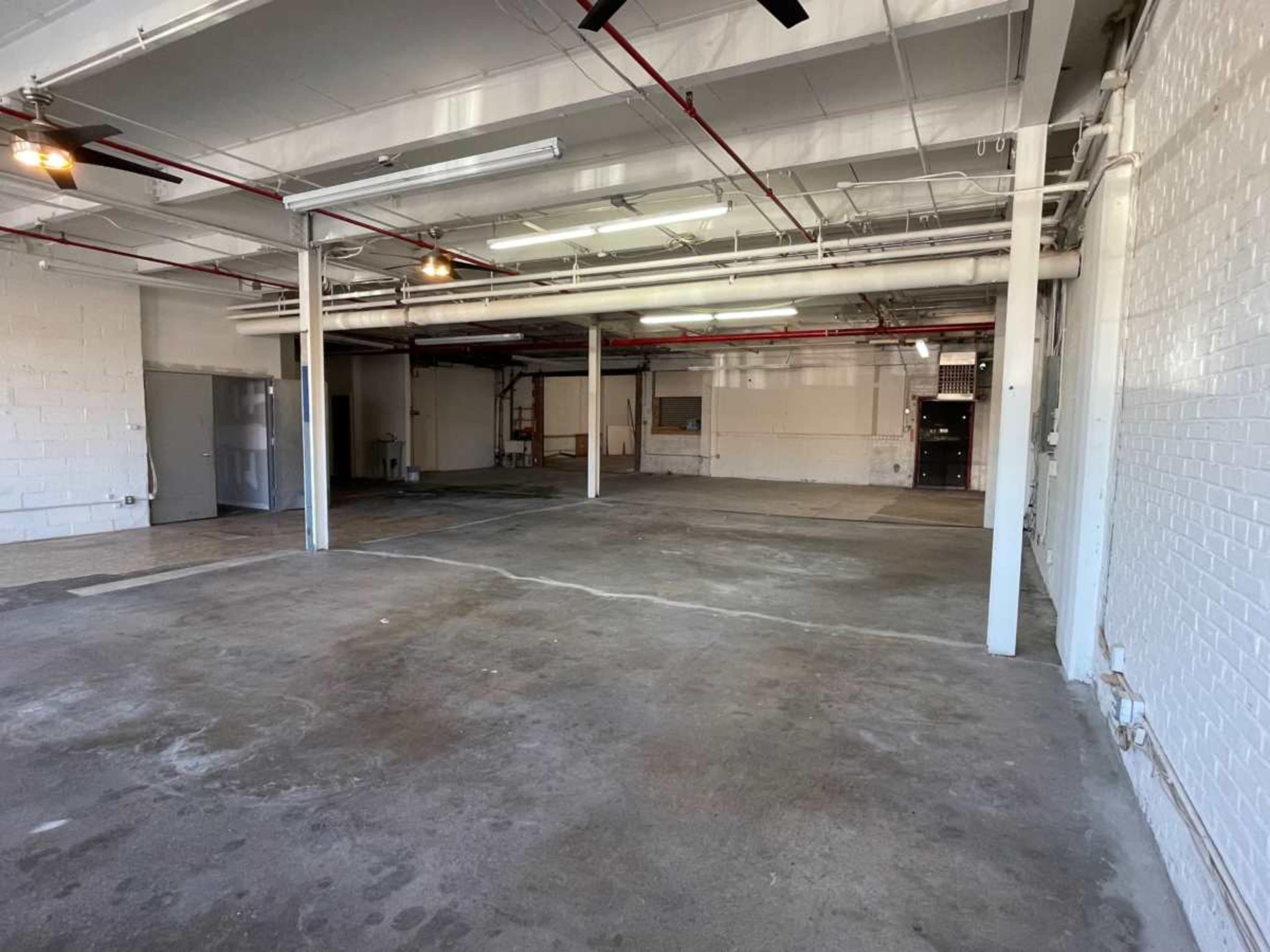 The image shows an empty, industrial-style interior space with concrete flooring and exposed pipes.