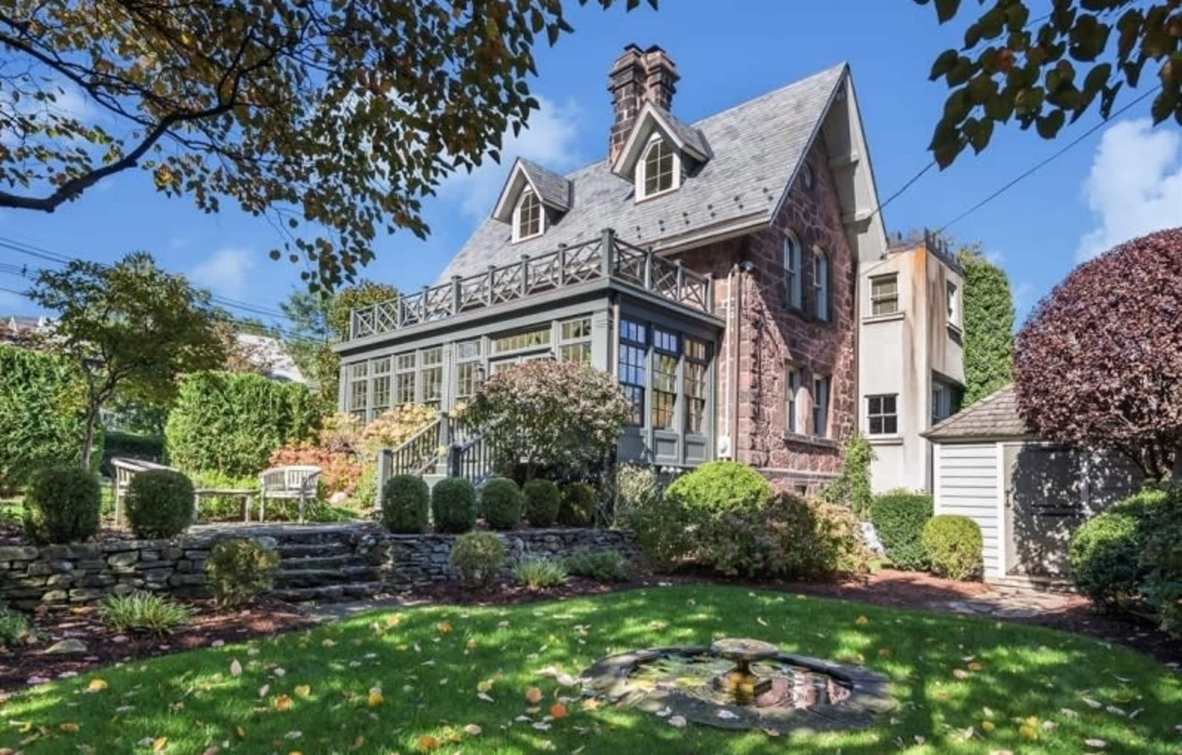 The image shows a well-maintained, historical-style house with a stone exterior, surrounded by a landscaped garden and a stone patio area.
