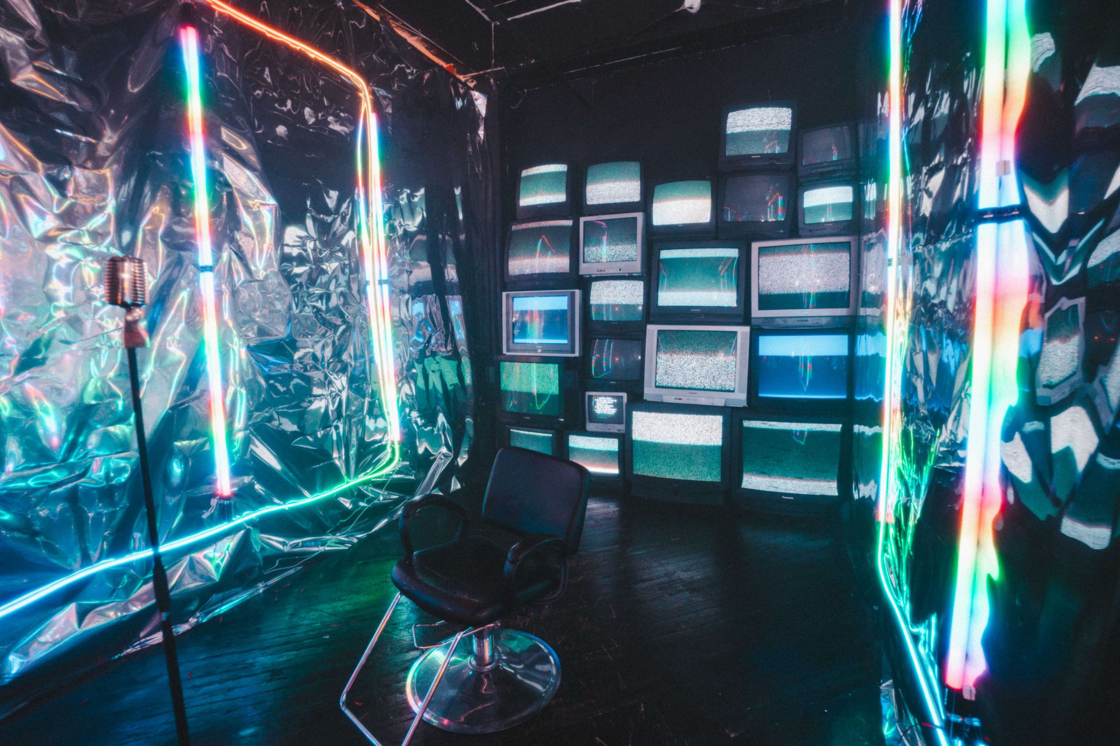 The image shows a reflective, metallic room with neon lights framing the walls and a row of vintage televisions displaying static on one side, alongside a swivel chair in the center.