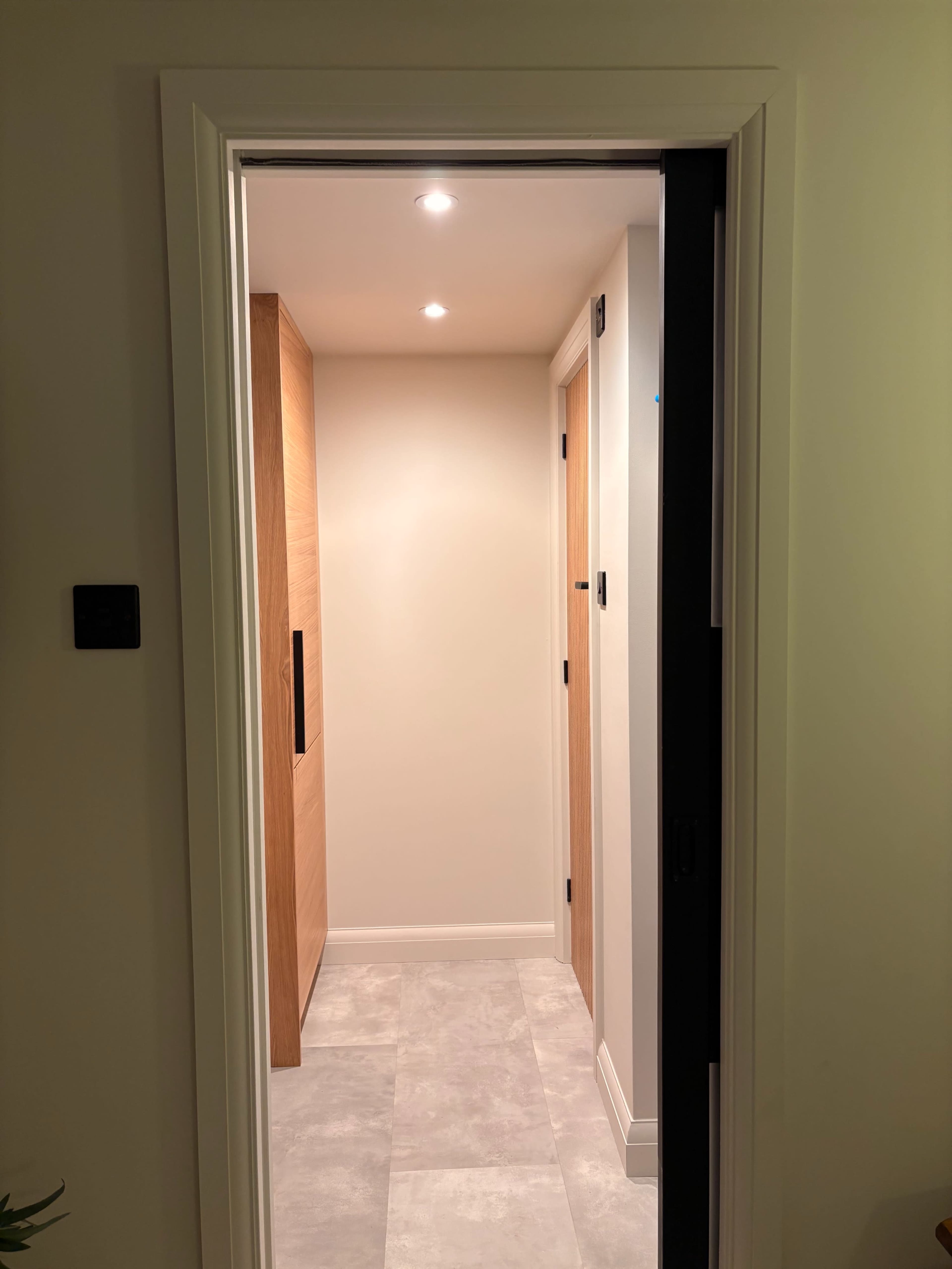 A narrow hallway with beige walls and a light-colored tile floor leads to a wooden door at the end.